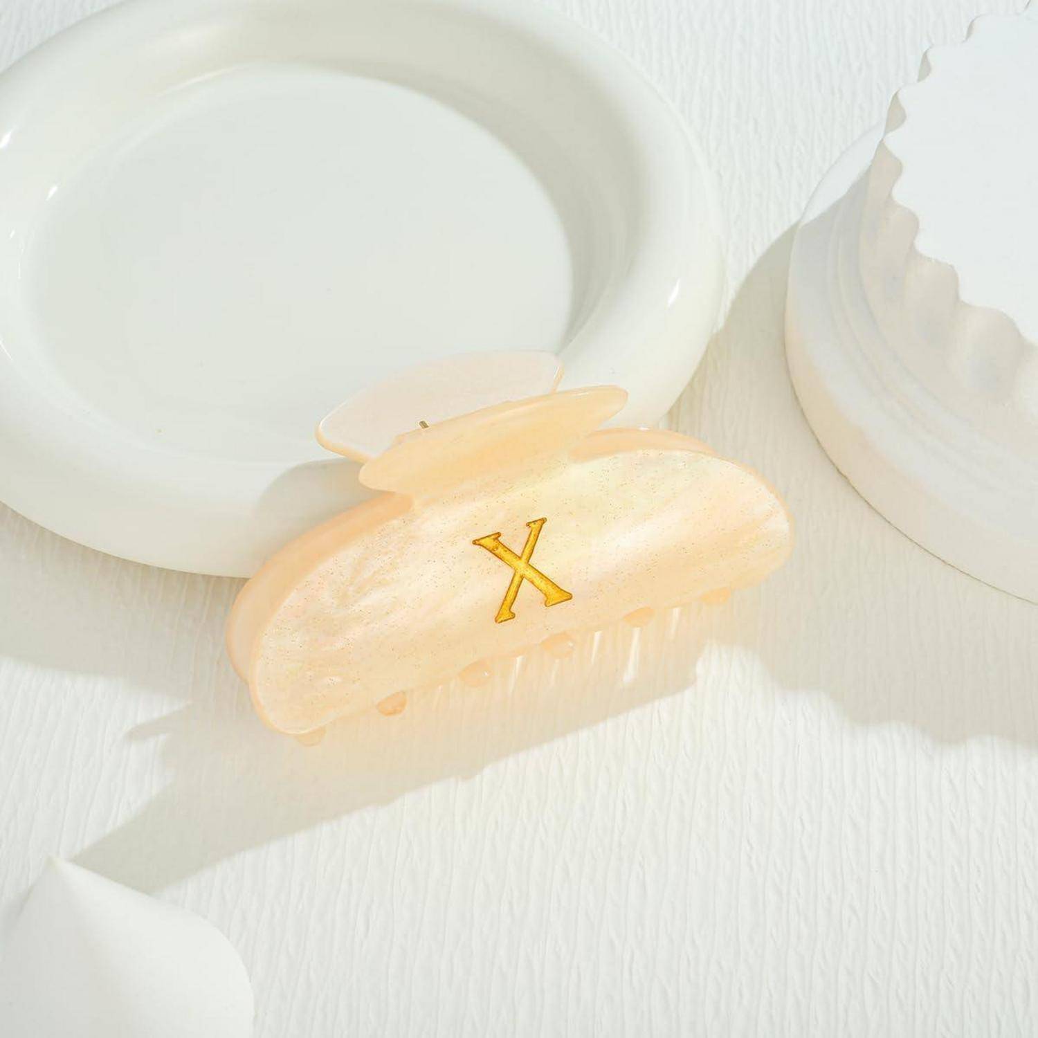 Ein pastellfarbener Haarclip mit einem goldenen 'X' ruht auf einem weißen Teller bei sanftem Licht und schafft eine minimalistische und elegante Komposition.
