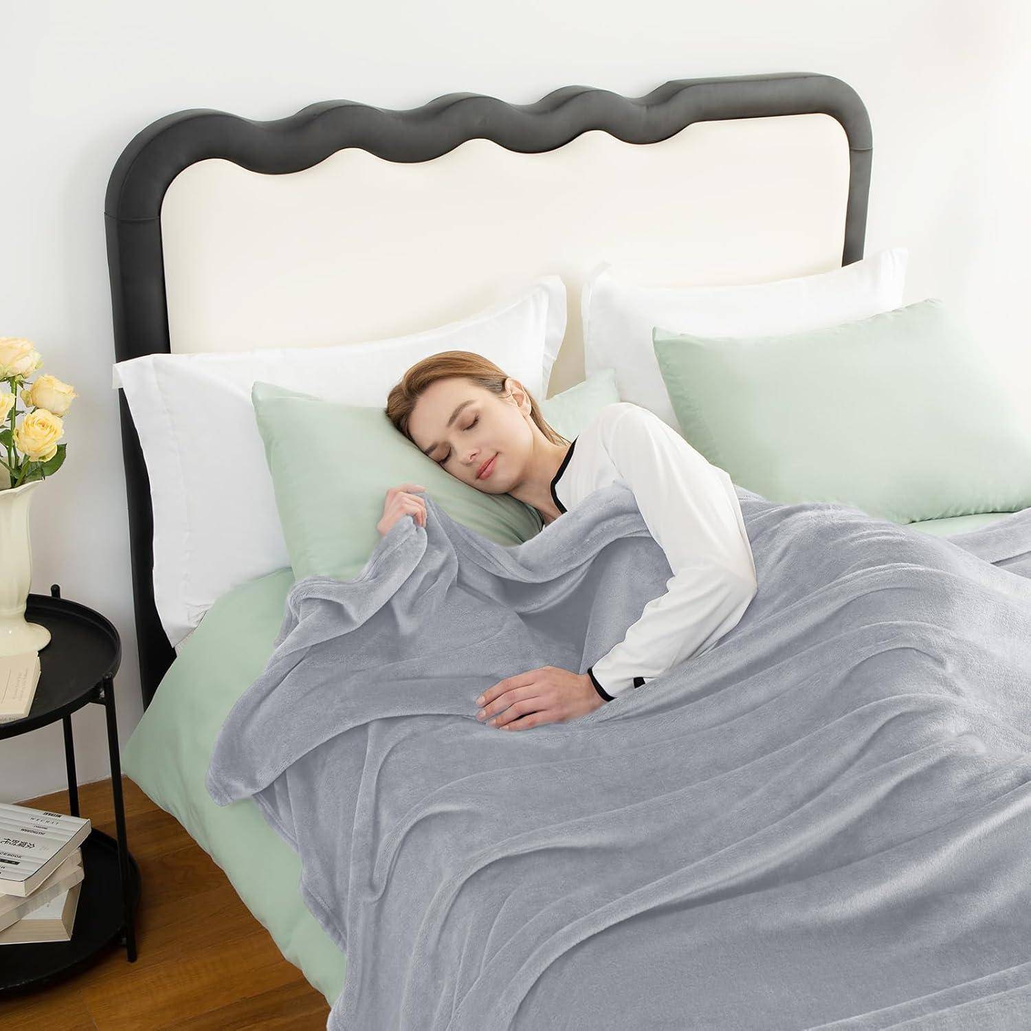 Eine Frau schläft friedlich in einem Bett, eingehüllt in eine graue Gewichtsdecke, umgeben von dekorativen Kissen und einem Nebentisch mit Blumen in der Nähe.