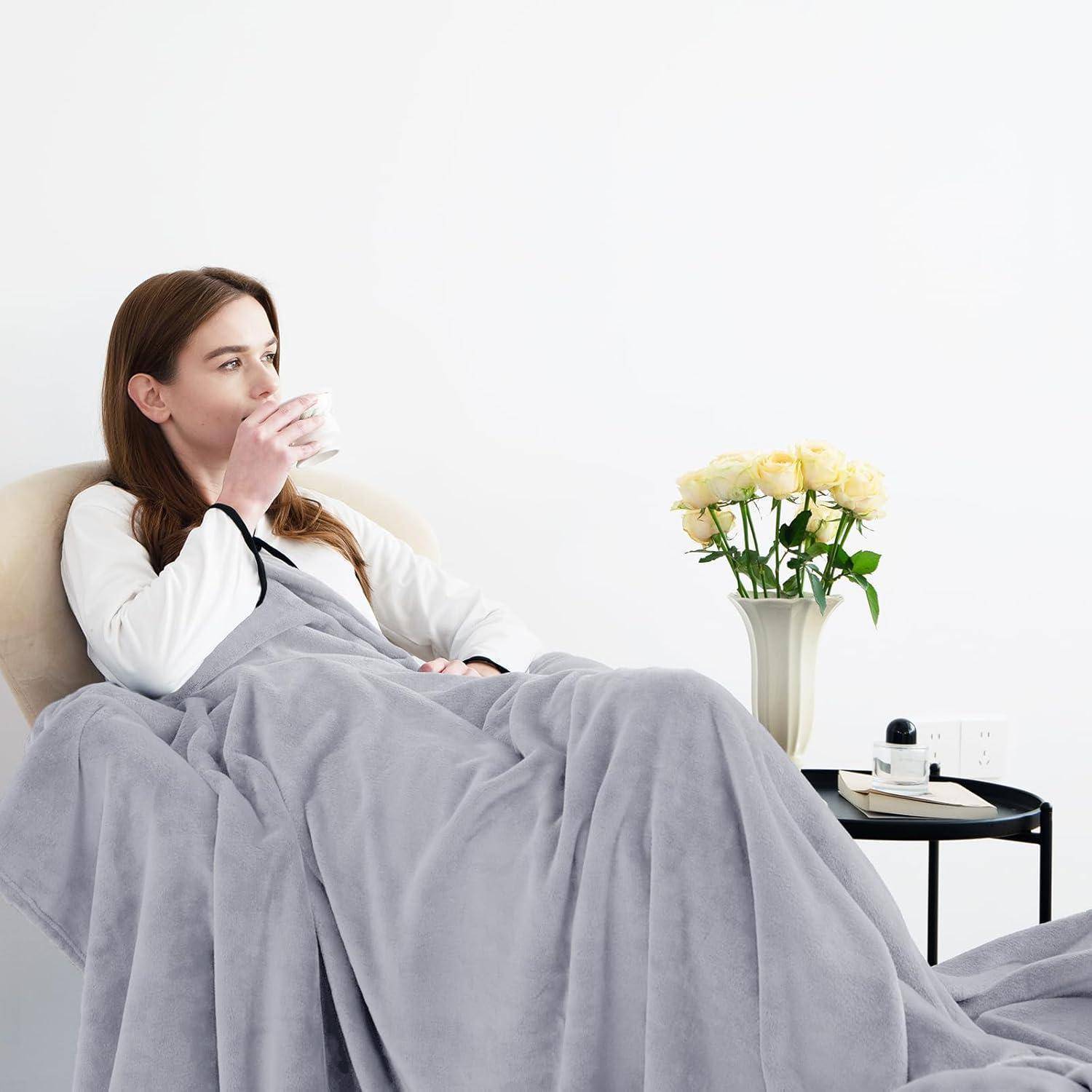 Eine Person sitzt auf einem Sofa, in eine graue Decke gehüllt, und nippt an einem Getränk. Eine Vase mit gelben Rosen und Büchern steht auf einem Tisch neben ihr.