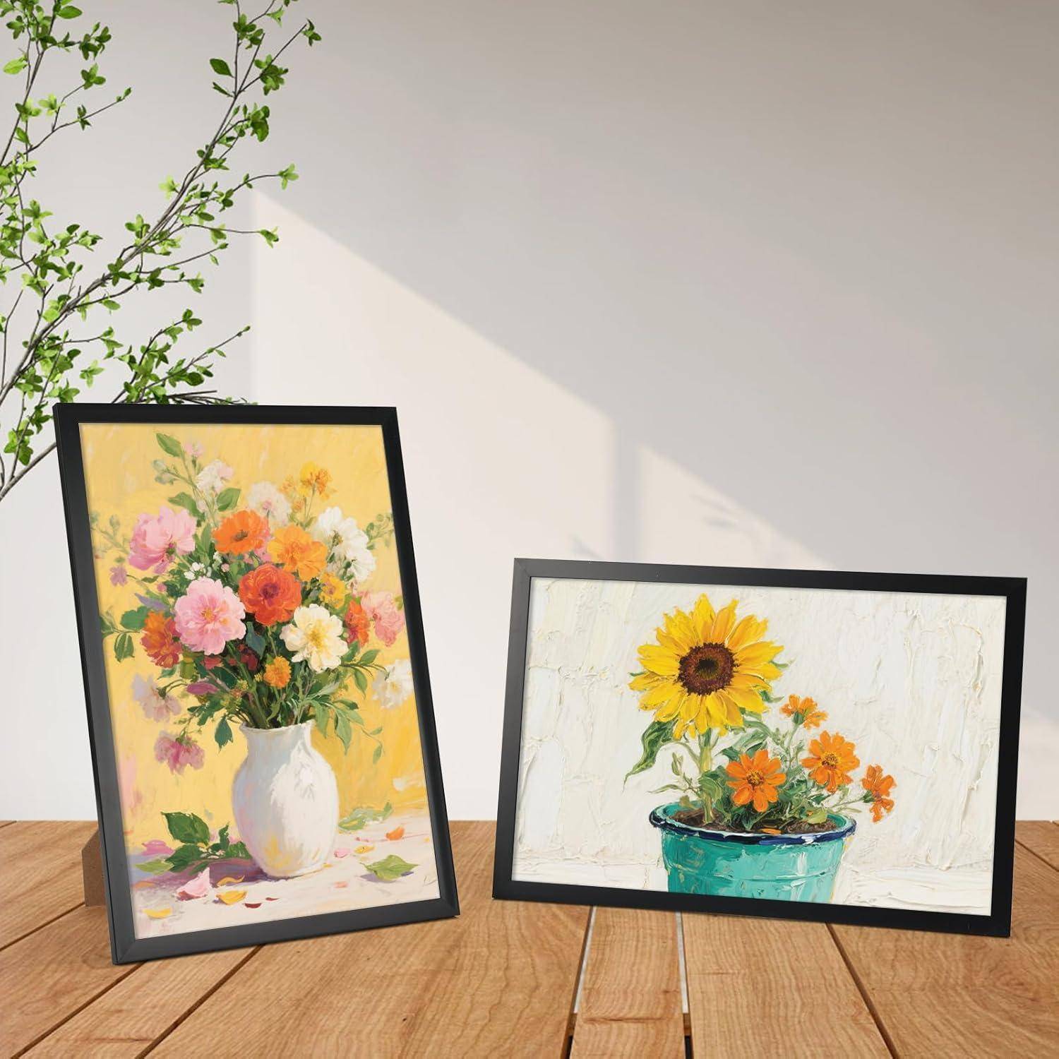 Zwei gerahmte Blumengemälde auf einem Holzboden: Das eine stellt eine Vase mit bunten Blumen dar, das andere zeigt Sonnenblumen und orangefarbene Blumen in einem Topf.