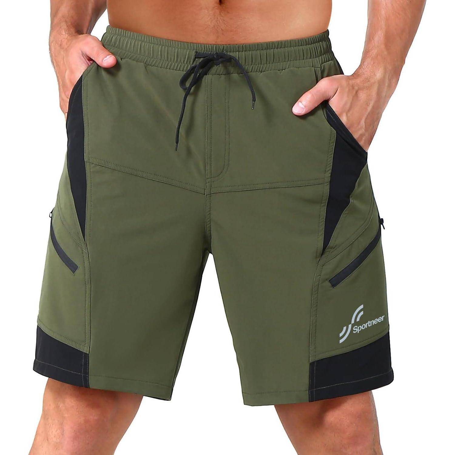 Eine Person, die olivgrüne Sportshorts mit schwarzen Seitenteilen trägt und vor einem einfarbigen Hintergrund steht. Das Markenlogo ist auf einem Bein sichtbar.