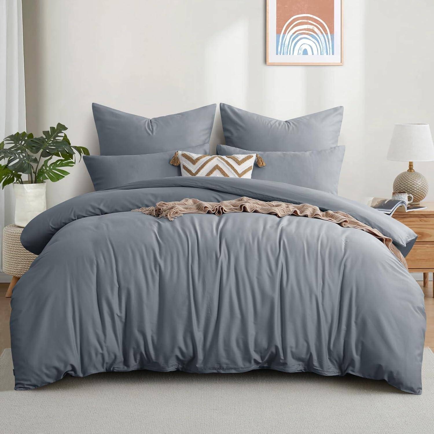 Ein sauber bezogenes Bett mit hellgrauer Bettwäsche, drei Kissen und einem dekorativen geometrisch gemusterten Kissen. Eine Pflanze und eine Lampe stehen auf einem Holznachttisch.
