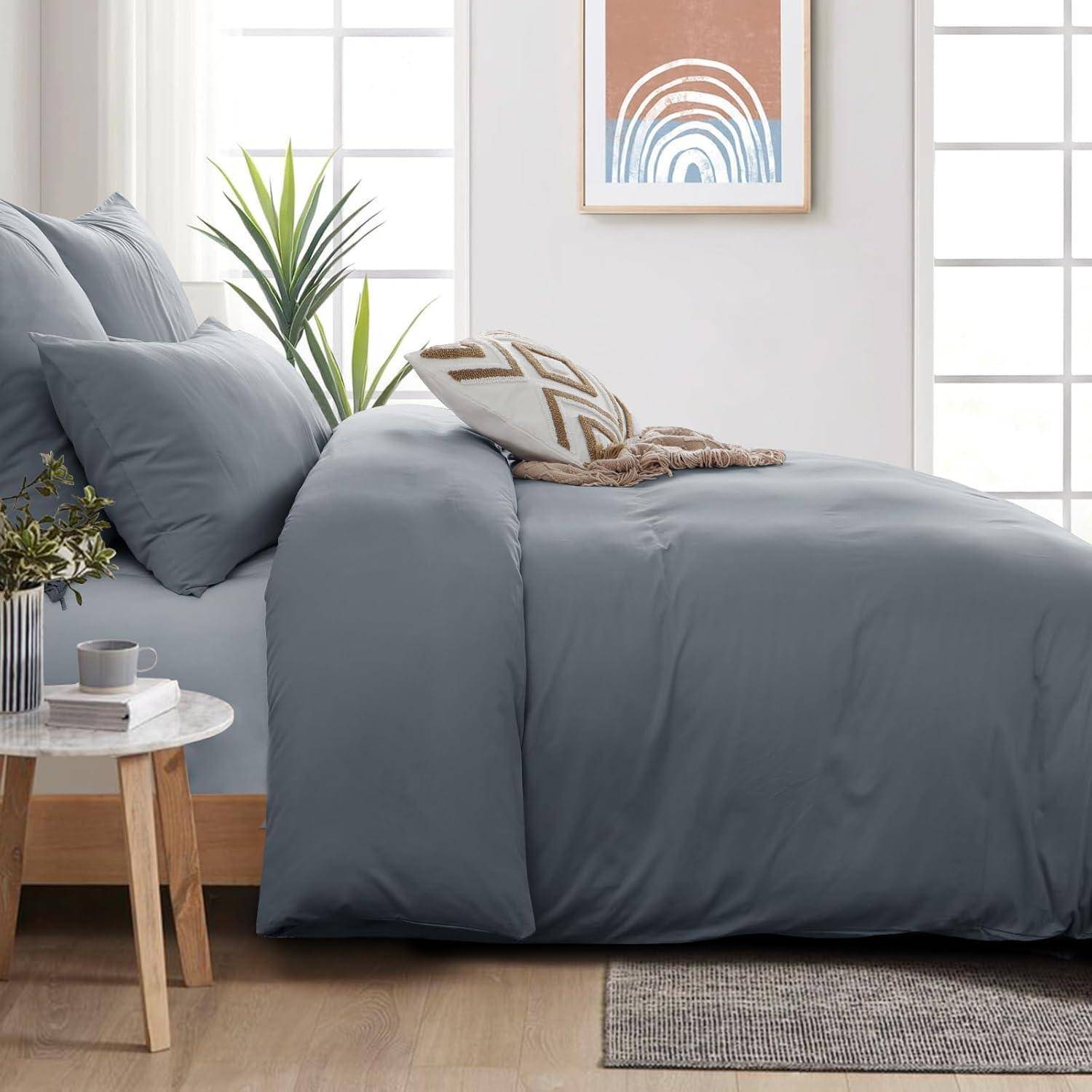 Ein ordentlich gemachtes Bett mit blauer Bettwäsche und gemusterten Kissen steht in einem Raum mit einem kleinen runden Beistelltisch, auf dem eine Pflanze und eine Tasse stehen. An der Wand hängt ein Kunstwerk.