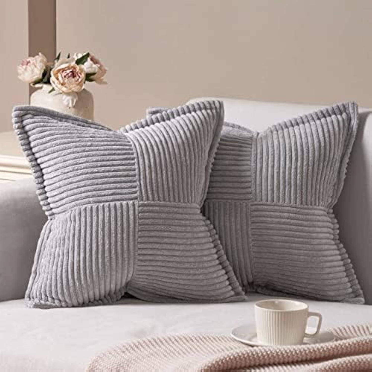 Zwei graue Cordkissen auf einem Sofa, mit einer weißen Tasse und Untertasse in der Nähe. Rosa Blumen in einer Vase sind im Hintergrund.