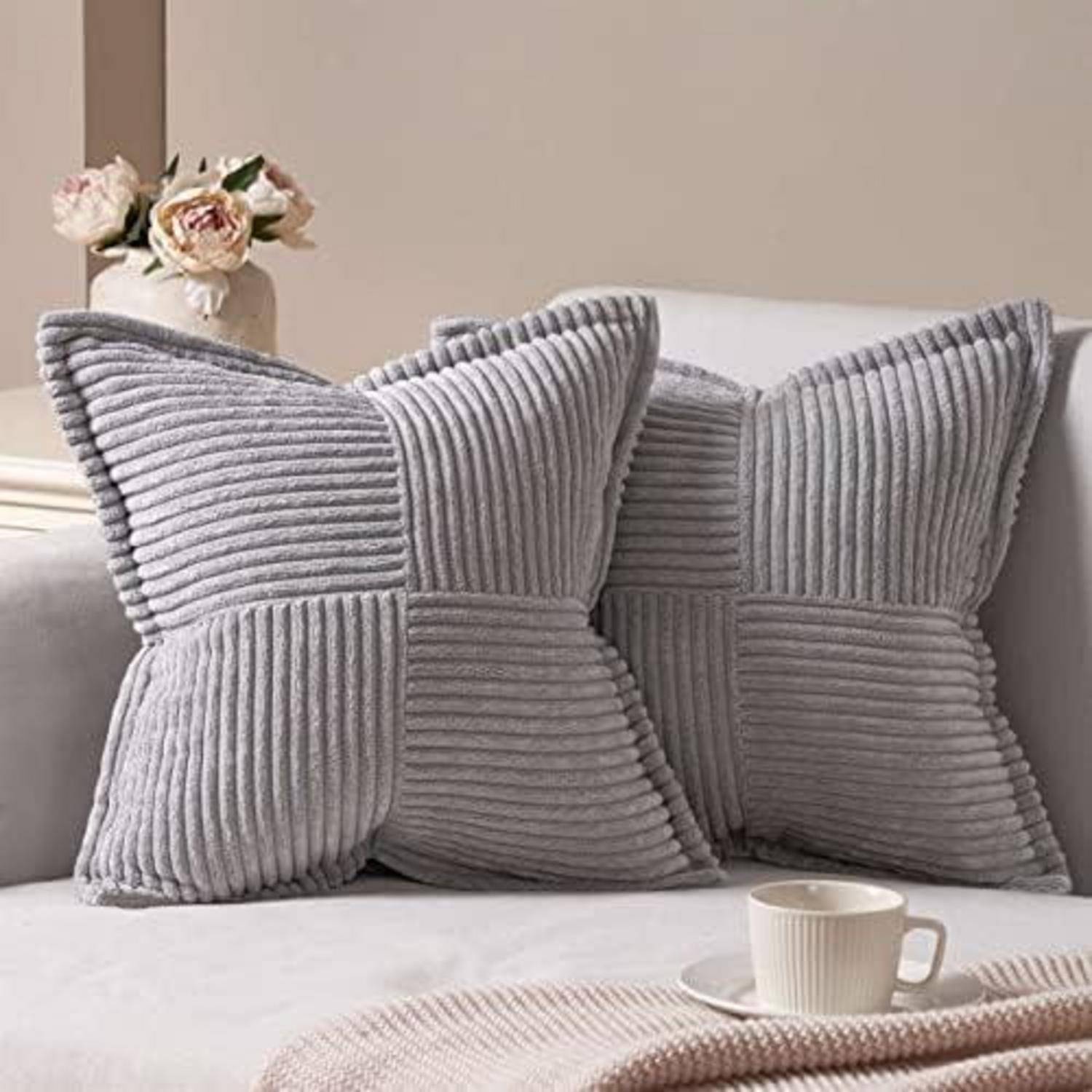 Zwei graue Cordkissen auf einem Sofa mit einer Kaffeetasse in der Nähe. Eine Vase mit rosa Blumen ist im Hintergrund. Eine gemütliche und einladende Atmosphäre.