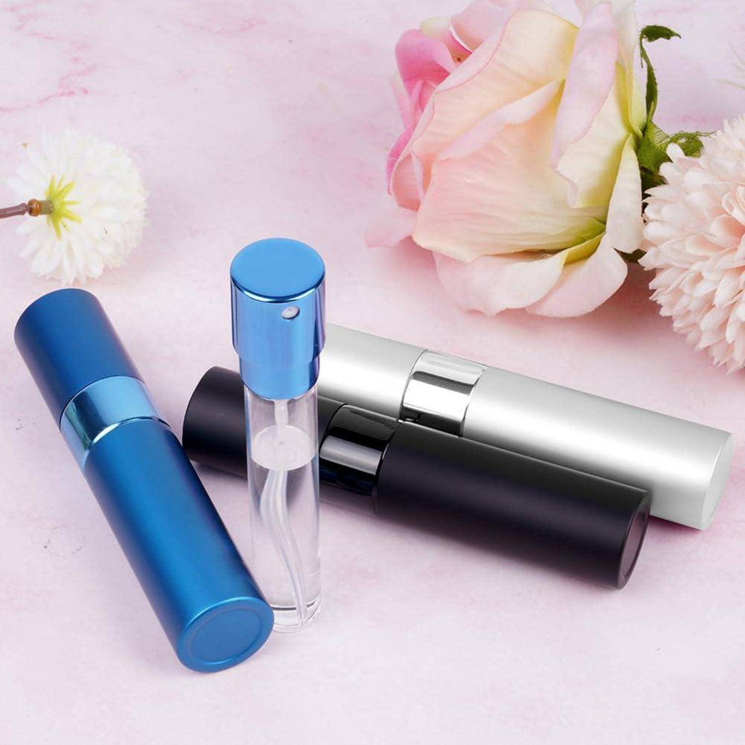 Tragbare Parfümflakons in Blau, Schwarz und Silber auf einer rosa Oberfläche mit Blumen im Hintergrund.