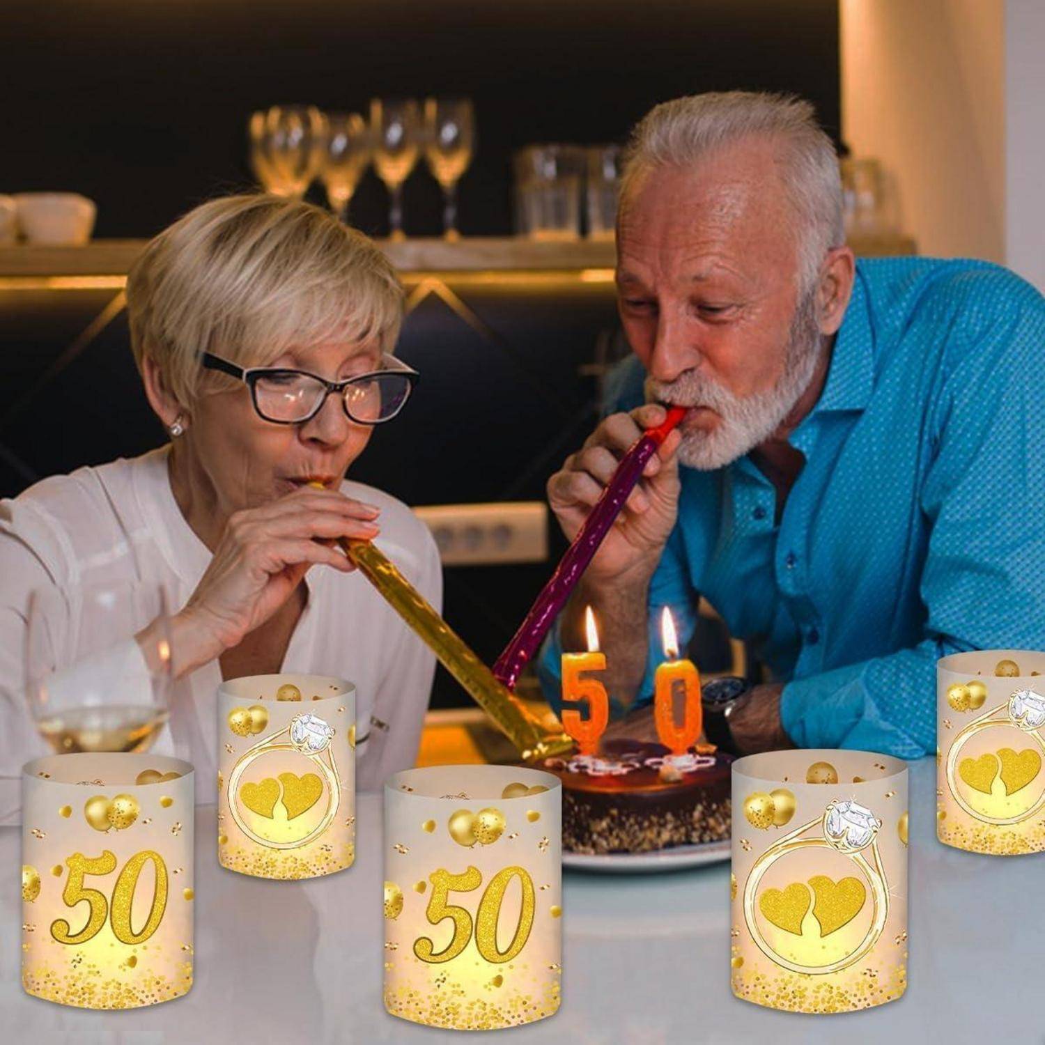 Ein älteres Ehepaar pustet die Kerzen auf einer '50' Geburtstagstorte aus. Der Tisch ist mit '50' thematischen Gegenständen dekoriert, was auf eine goldene Hochzeitsfeier hinweist.