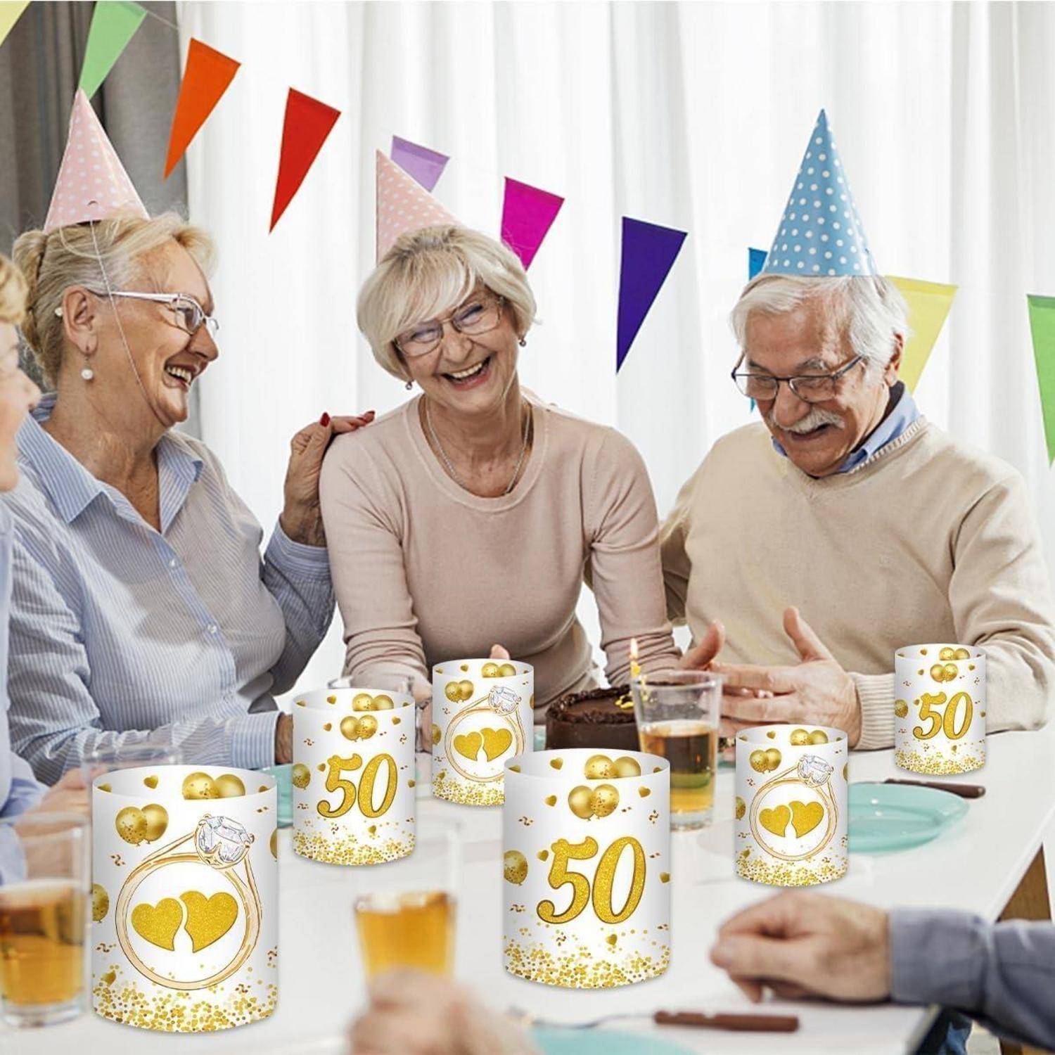 Eine Gruppe älterer Menschen mit Partyhüten sitzt um einen Tisch mit Getränken und feiert eine 50-jährige Jubiläumsfeier. Dekorierte Tassen und Partydekoration sind sichtbar.