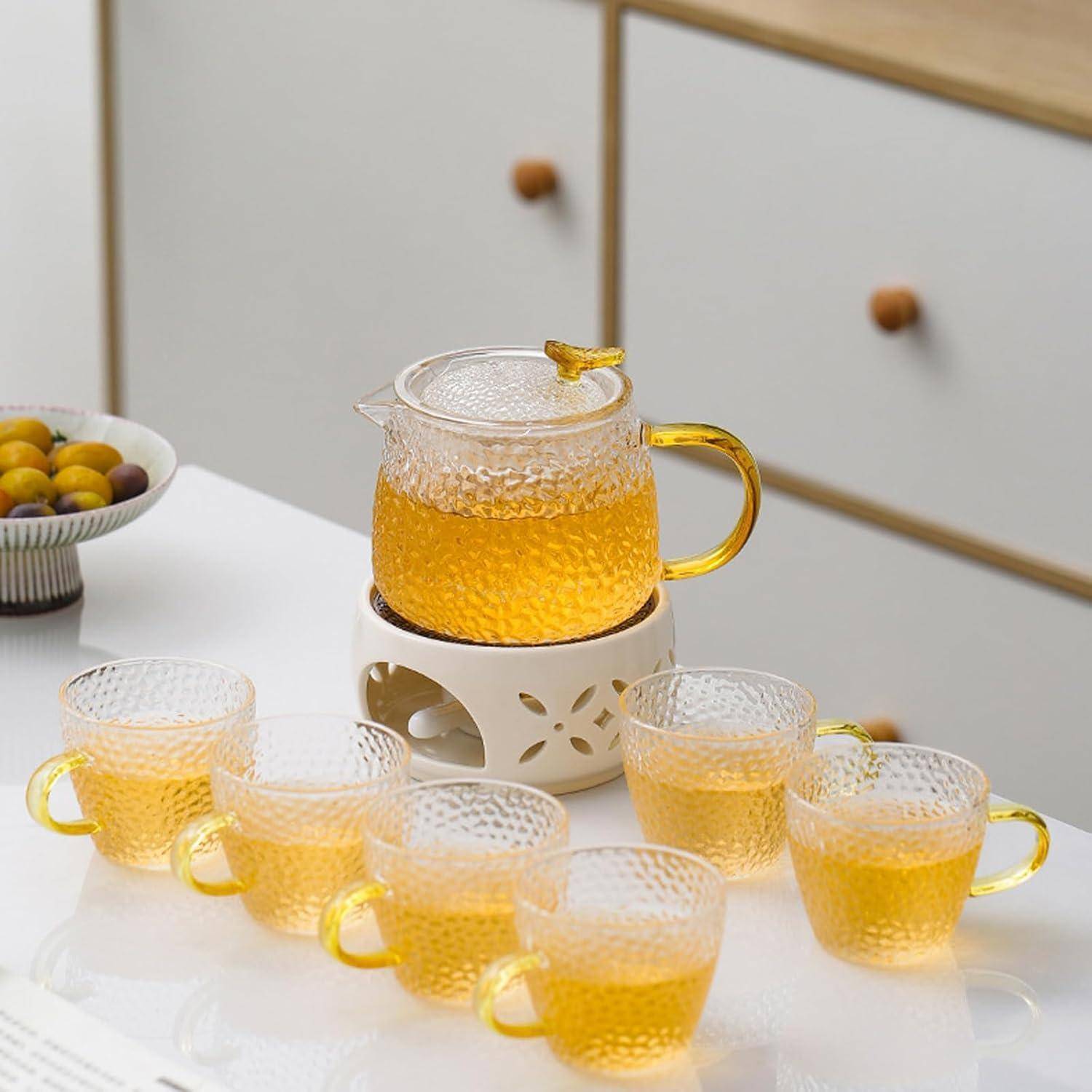 Ein Glasteekannen mit gelbem Tee auf einem Rechaud, umgeben von sechs passenden Tassen auf einem weißen Tisch. Eine Schüssel mit Pflaumen befindet sich im Hintergrund.