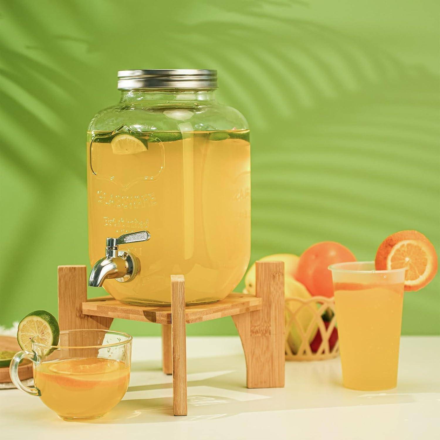 Glasspender gefüllt mit Limonade auf einem Holzständer, daneben ein Glas und eine Tasse, mit Zitrusfrüchten im Hintergrund vor einem grünen Hintergrund.