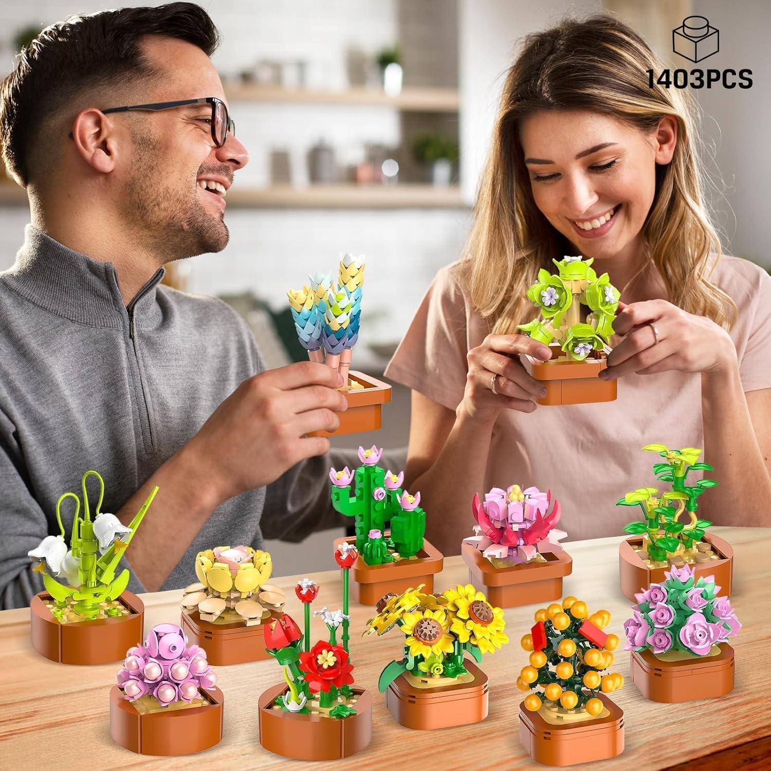 Zwei Personen lächeln, während sie verschiedene bunte Topfpflanzenmodelle auf einem Tisch zusammenbauen. Die Modelle zeigen Blumen und Sukkulenten.
