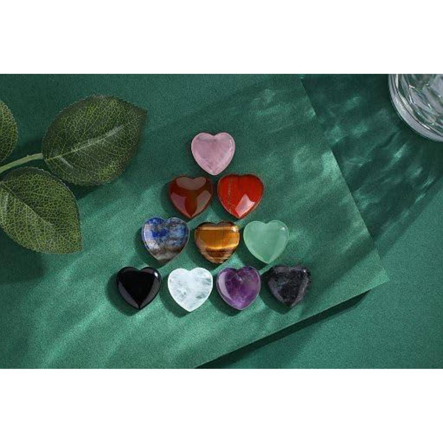 Eine Gruppe von zwölf herzförmigen Steinen, die auf einer grünen Oberfläche zu einem Dreieck angeordnet sind, wobei jeder Stein eine andere Farbe hat, neben grünen Blättern.