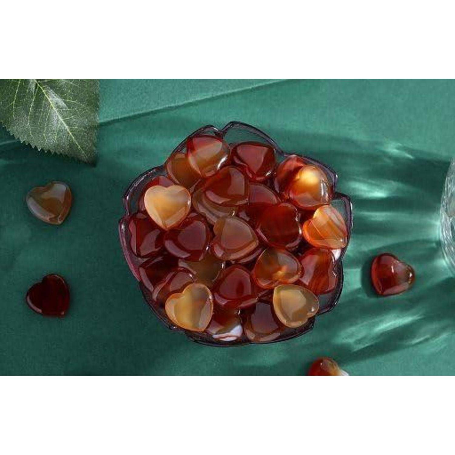 Eine Schüssel gefüllt mit herzförmigen Gummibonbons in roten und orangefarbenen Tönen auf einer grünen Oberfläche. Eine teilweise sichtbare Ecke einer blättrigen Pflanze ist im Bild.