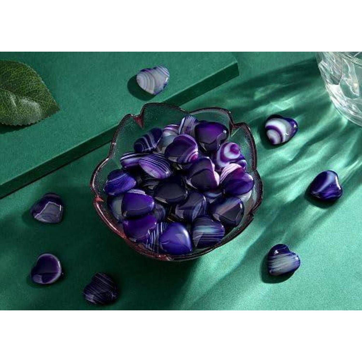 Eine Glasschüssel, gefüllt mit polierten violetten herzförmigen Steinen, ist von ähnlichen Steinen auf einer grünen Oberfläche umgeben. Ein Blatt und ein Kristallglas befinden sich in der Nähe.