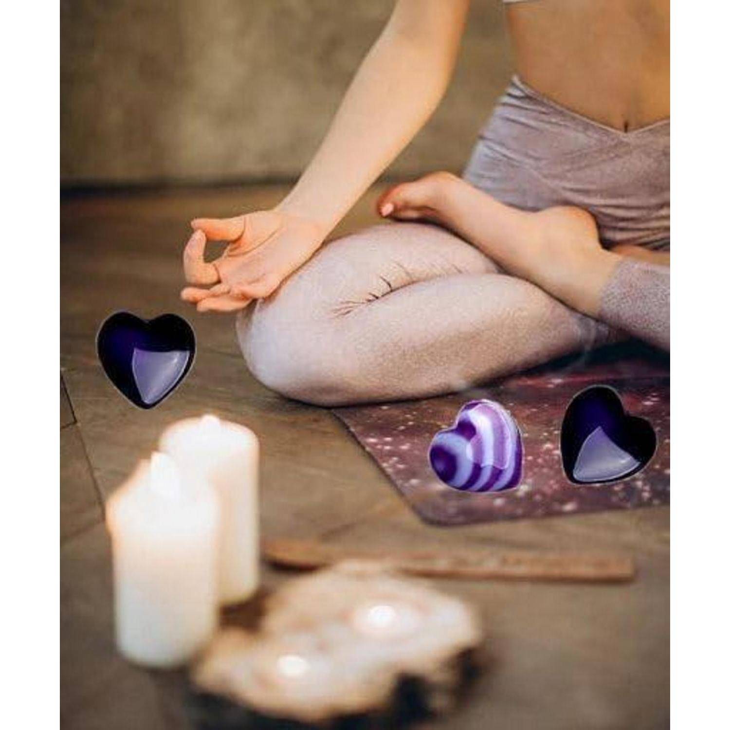 Eine Person in einer sitzenden Yoga-Pose mit gekreuzten Beinen und offener Handfläche in der Nähe von Kerzen und herzförmigen Objekten; eine ruhige, meditative Umgebung.