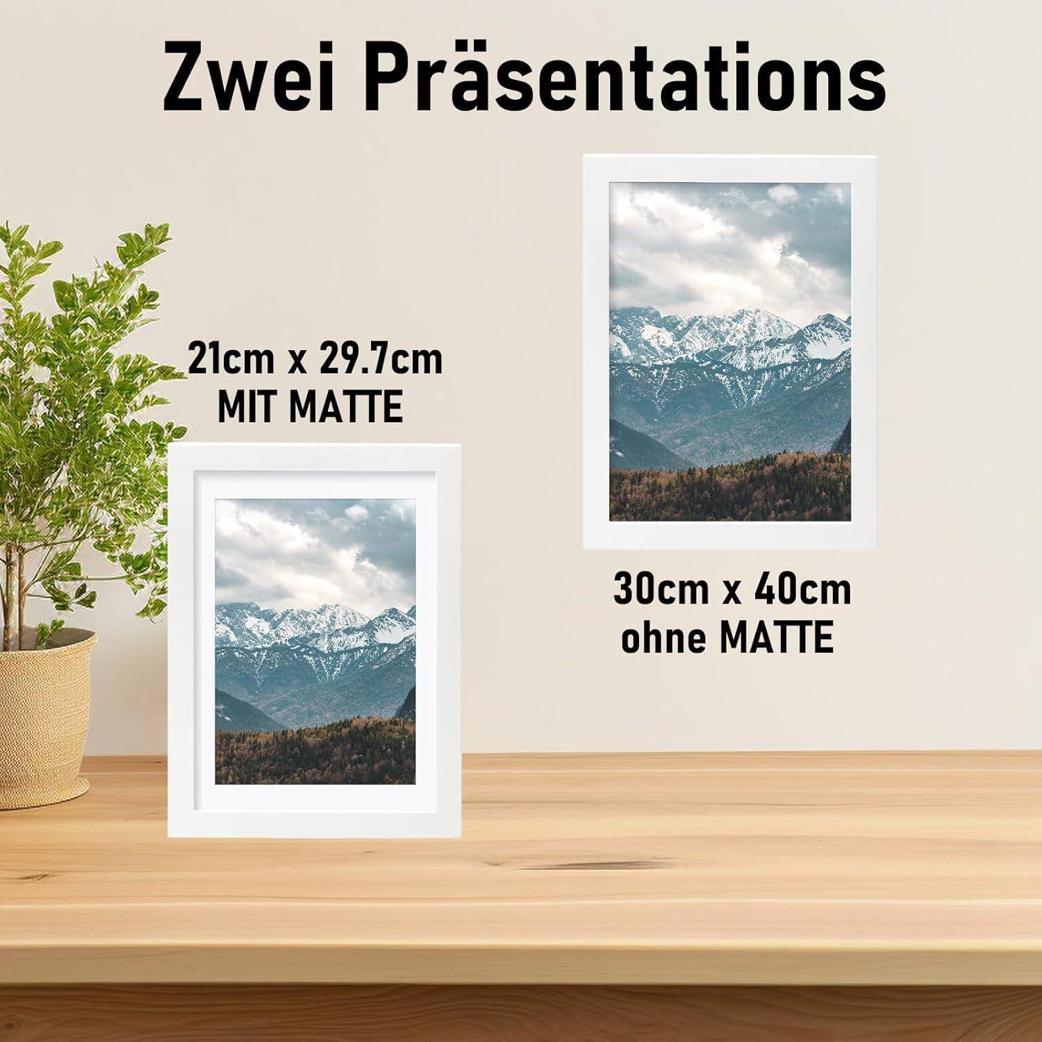 Ein weißer Rahmen mit einem Bergbild steht auf einem Tisch neben einer Pflanze. Text oben: 'Zwei Präsentationen'. Beschreibungen: '21cm x 29,7cm MIT MATTE' und '30cm x 40cm ohne MATTE'.