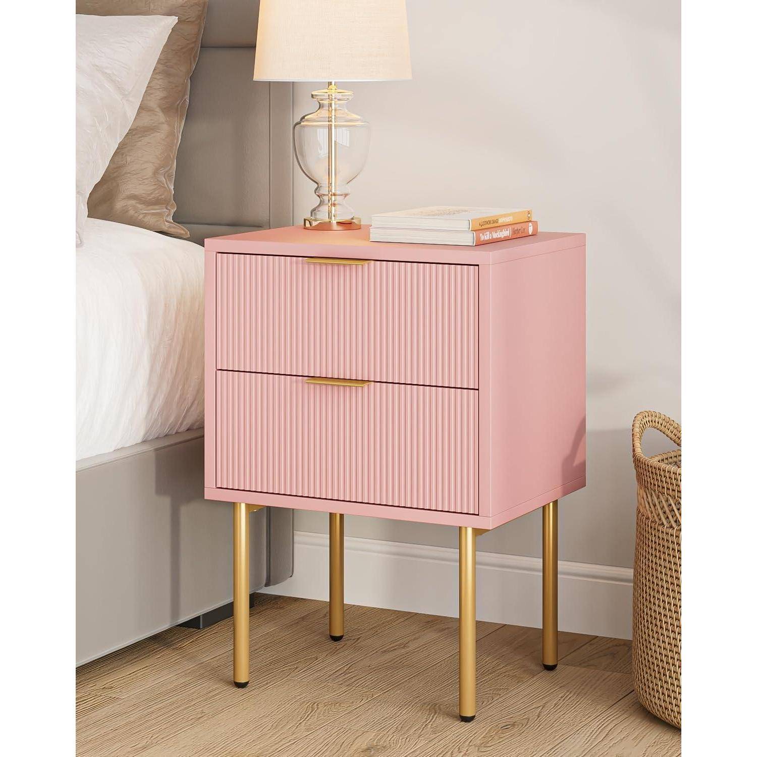 Ein rosa Nachttisch mit zwei Schubladen steht neben einem Bett. Er hat goldene Beine und eine Lampe oben drauf, begleitet von einem Buch und einem kleinen Dekorationsobjekt.