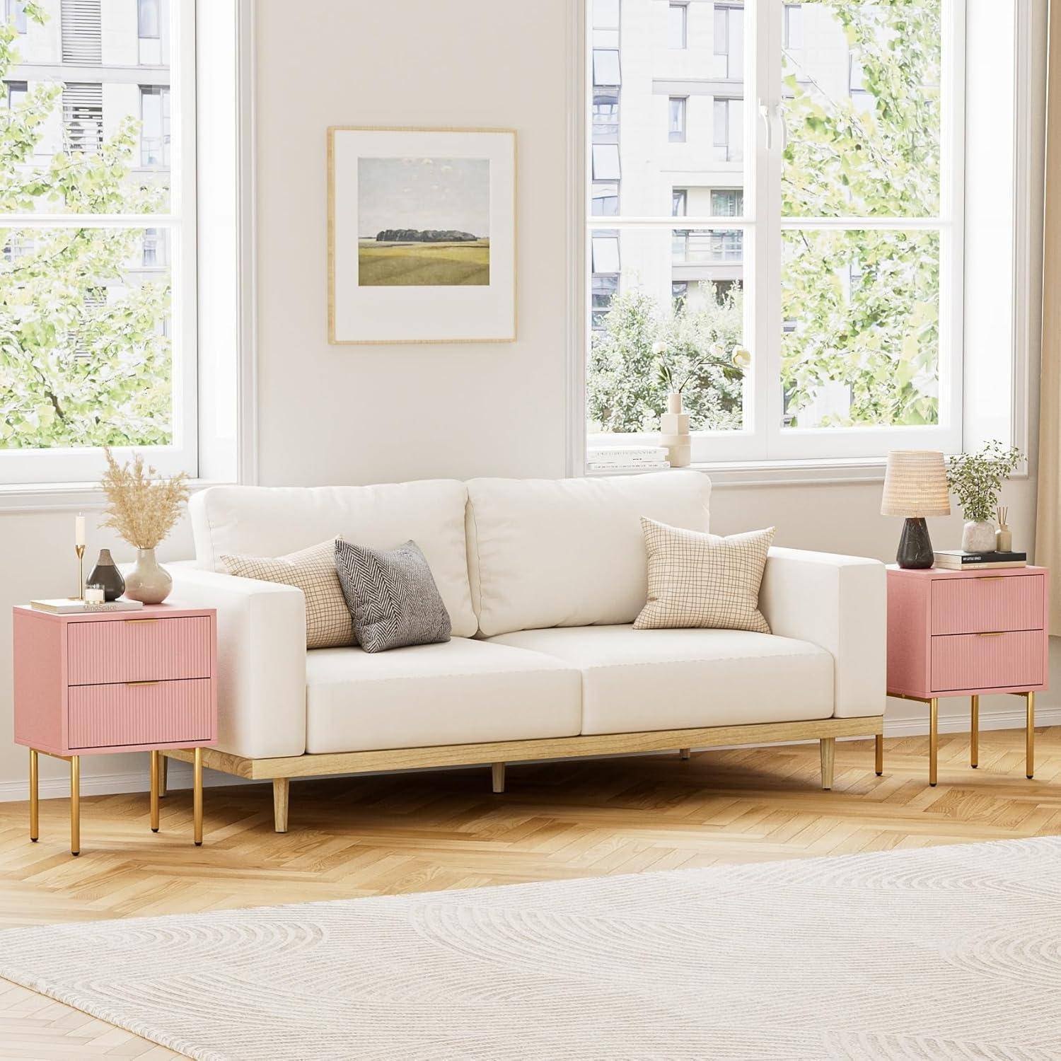 Ein modernes Wohnzimmer mit einem weißen Sofa, rosa Beistelltischen und einem beigen Teppich. Große Fenster zeigen grüne Bäume draußen. Ein gerahmtes Bild hängt über dem Sofa.
