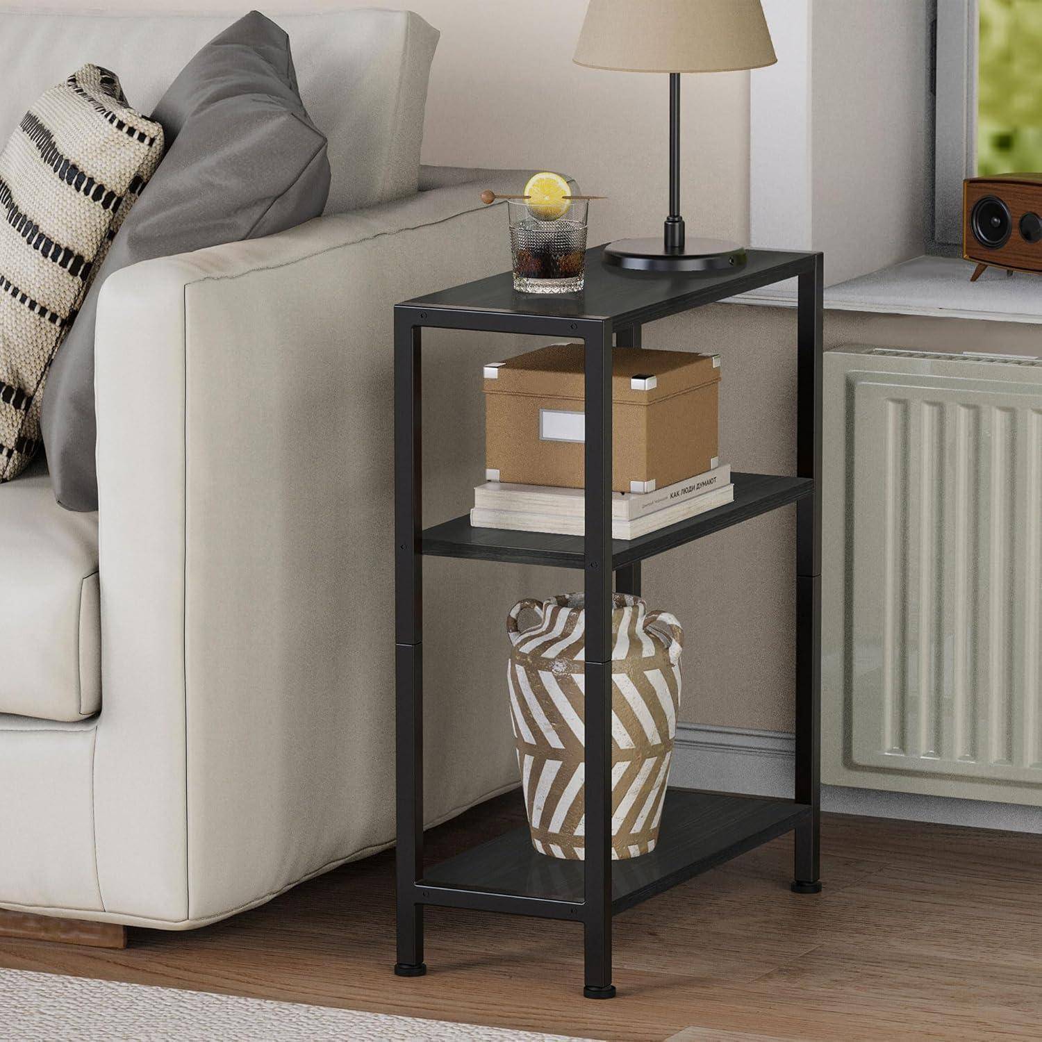 Ein schwarzer Beistelltisch mit drei Ablagen steht neben einem hellgrauen Sofa. Auf den Ablagen befinden sich ein Getränk, eine Lampe, Bücher und eine Ziervase.