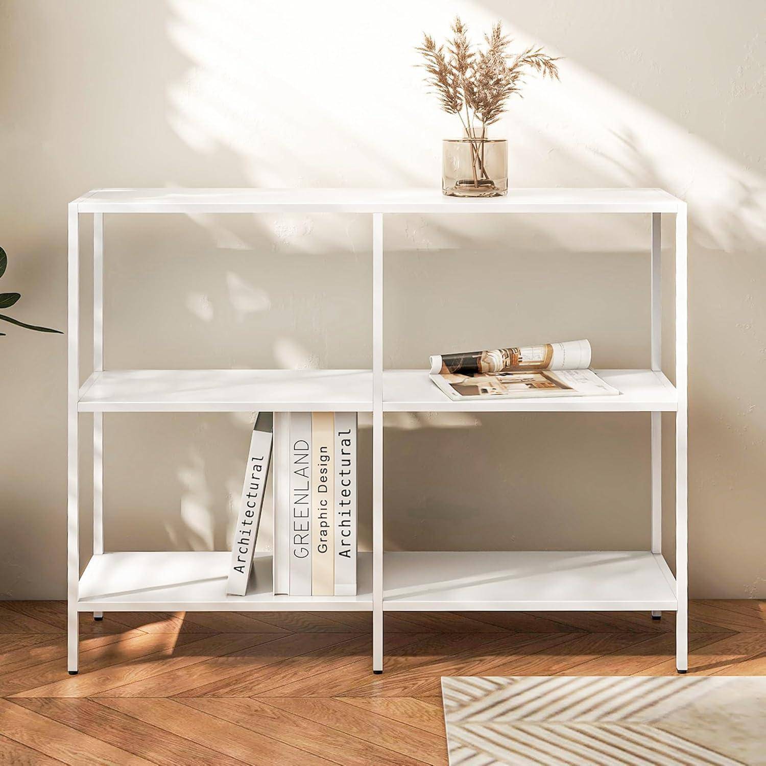 Ein minimalistisches weißes Regal mit drei Ebenen, auf dem mehrere Bücher und ein zusammengerolltes Tuch liegen. Eine kleine Vase mit Pflanzen steht oben, in einem hell beleuchteten Raum.