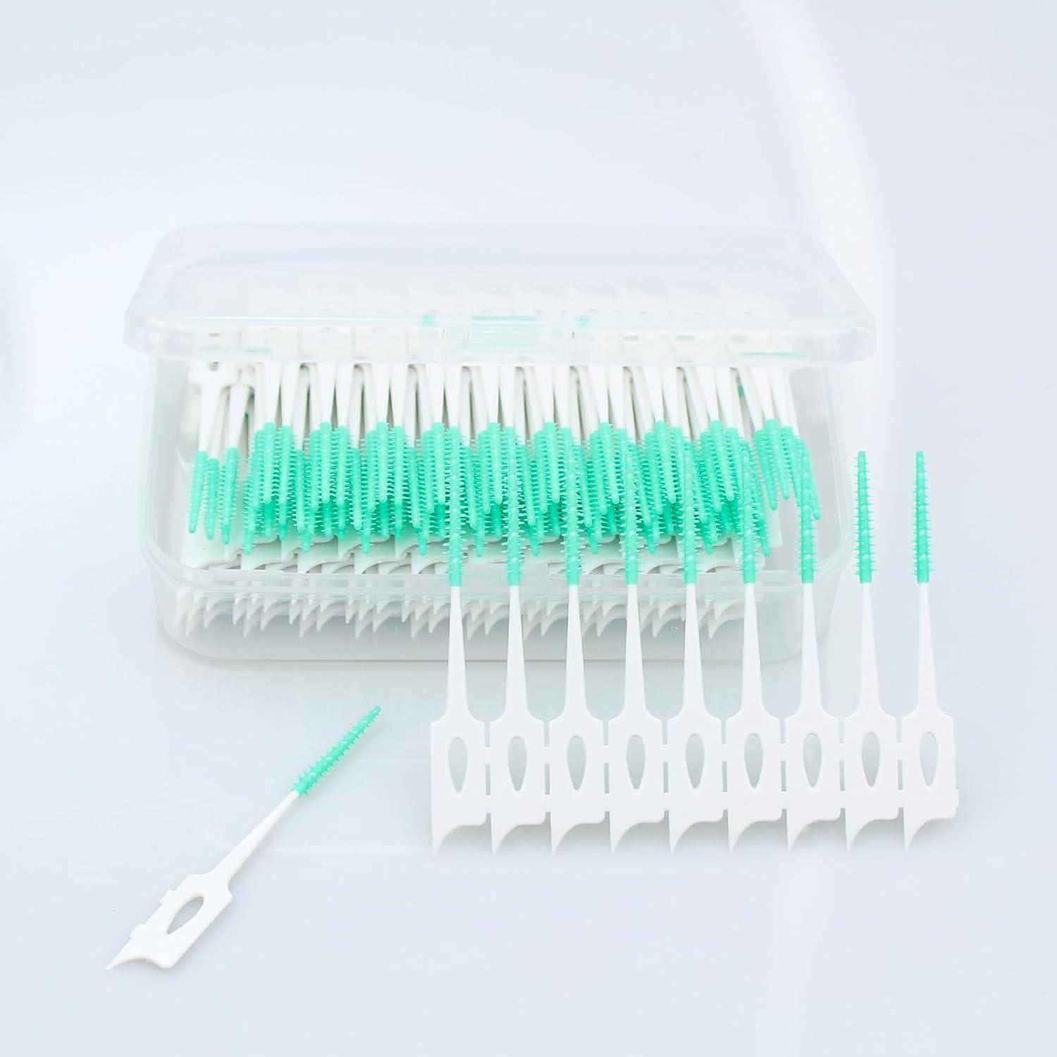 Eine durchsichtige Plastikbox mit weißen und grünen Interdentalbürsten, wobei eine Bürste flach davor liegt.