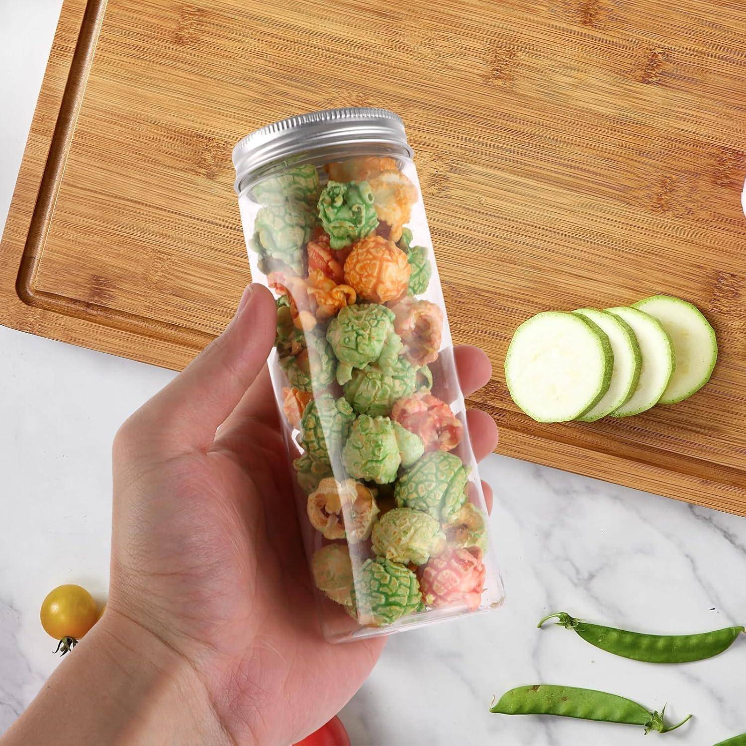 Eine Hand hält ein durchsichtiges Glas gefüllt mit buntem Popcorn auf einer Marmoroberfläche. In der Nähe liegen geschnittene Gurke, Zuckerschoten und ein Holzbrett.
