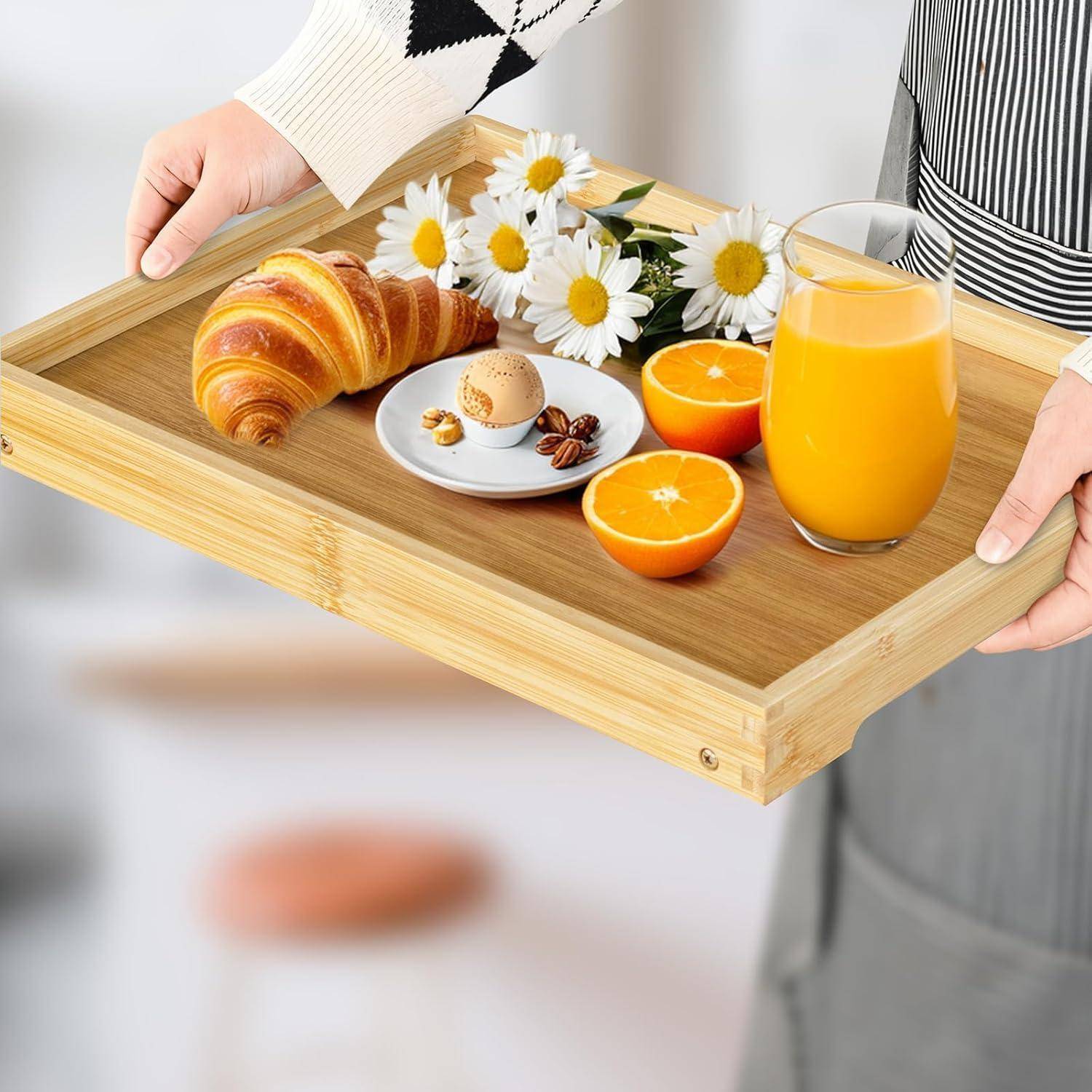 Eine Person hält ein Holztablett mit einem Croissant, Eiern, Orangenscheiben, Gänseblümchen und einem Glas Orangensaft, was einen Frühstückskontext andeutet.