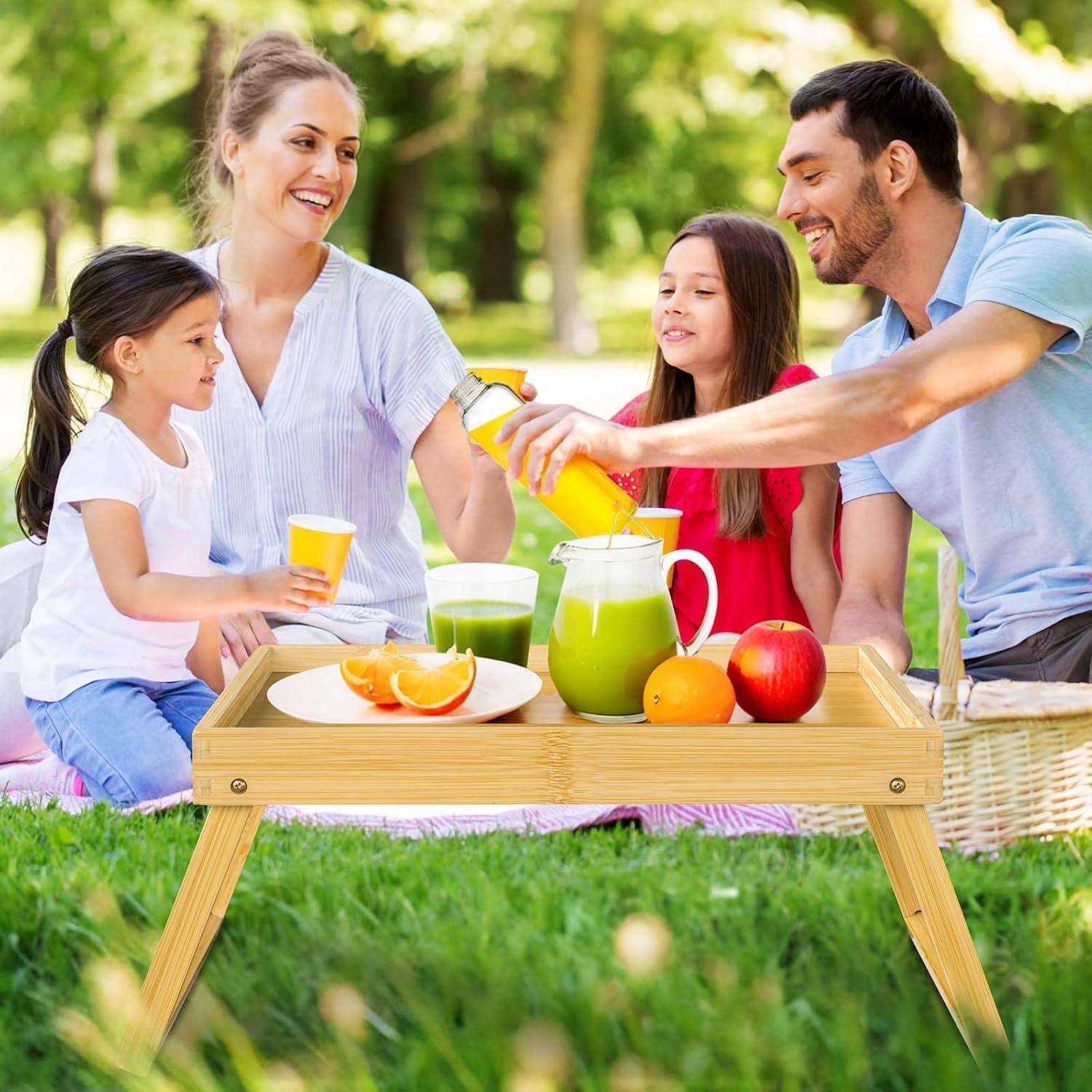 Eine Familie genießt ein Picknick auf einer Wiese, mit einem kleinen Tisch, auf dem Saft, Äpfel und Brot stehen. Eine Frau schenkt Saft ein, während sie mit anderen spricht.
