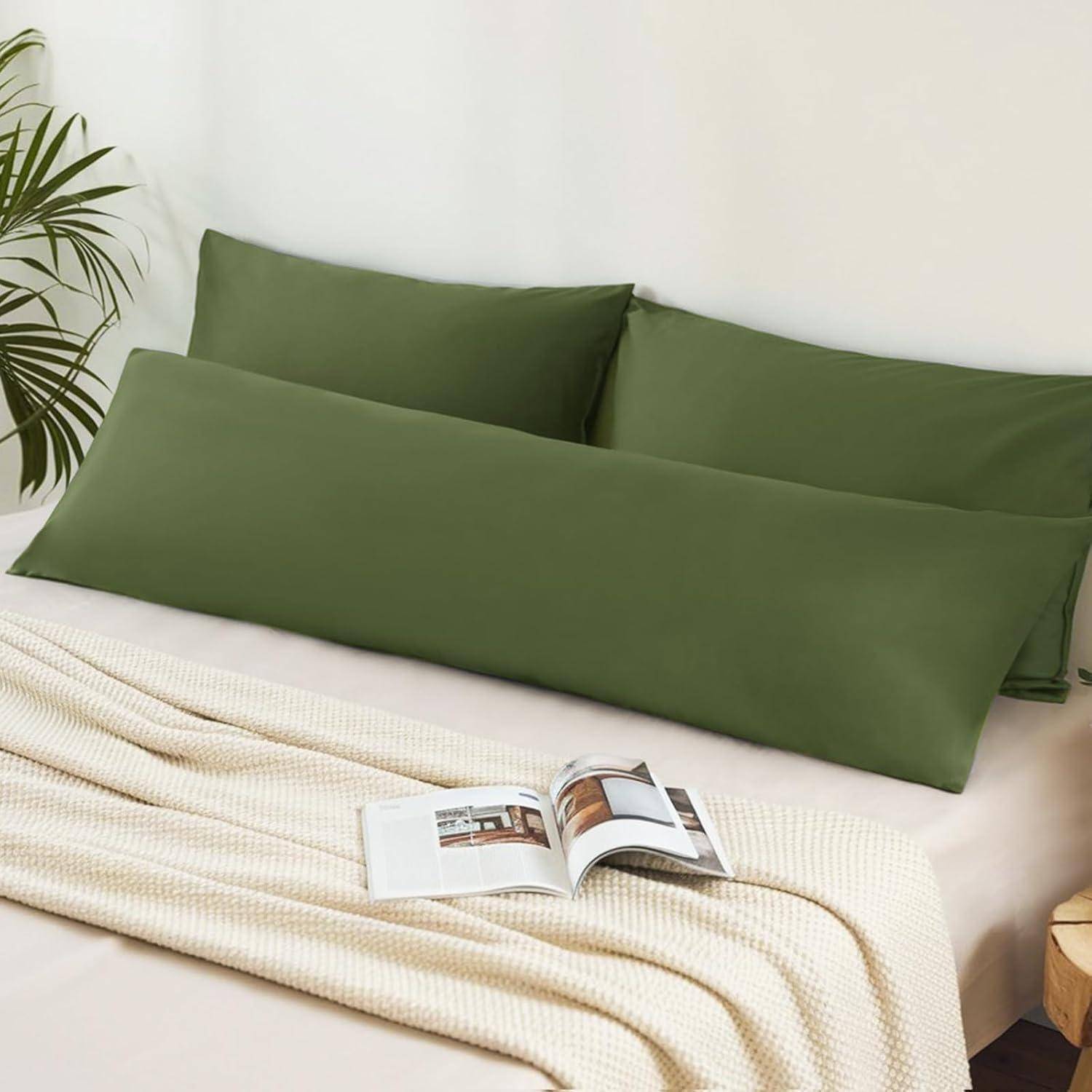 Ein Bett mit grünen Kissen und einer cremefarbenen Decke; ein aufgeschlagenes Buch liegt auf dem Bett. Eine kleine Pflanze steht in der Ecke des Zimmers.