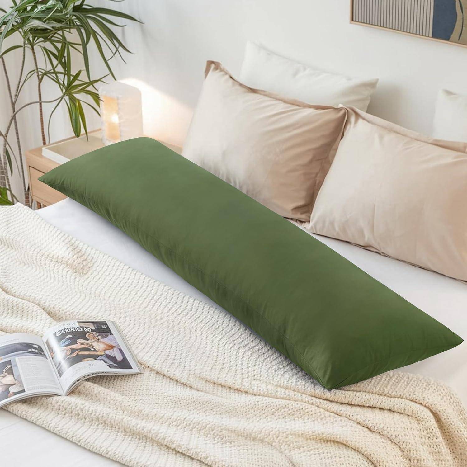 Ein langer grüner Kissenbezug liegt auf einem sorgfältig gemachten Bett mit beigen Kissen und einer weißen Decke. Ein aufgeschlagenes Magazin und eine Topfpflanze befinden sich in der Nähe.