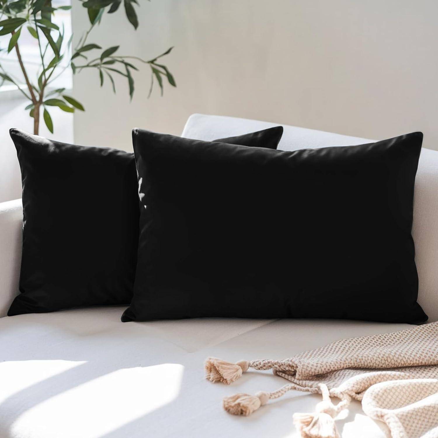 Zwei schwarze Kissen liegen auf einem weißen Sofa, im Hintergrund eine blattreiche Pflanze. Sonnenlicht wirft sanfte Schatten und schafft eine gemütliche Atmosphäre.