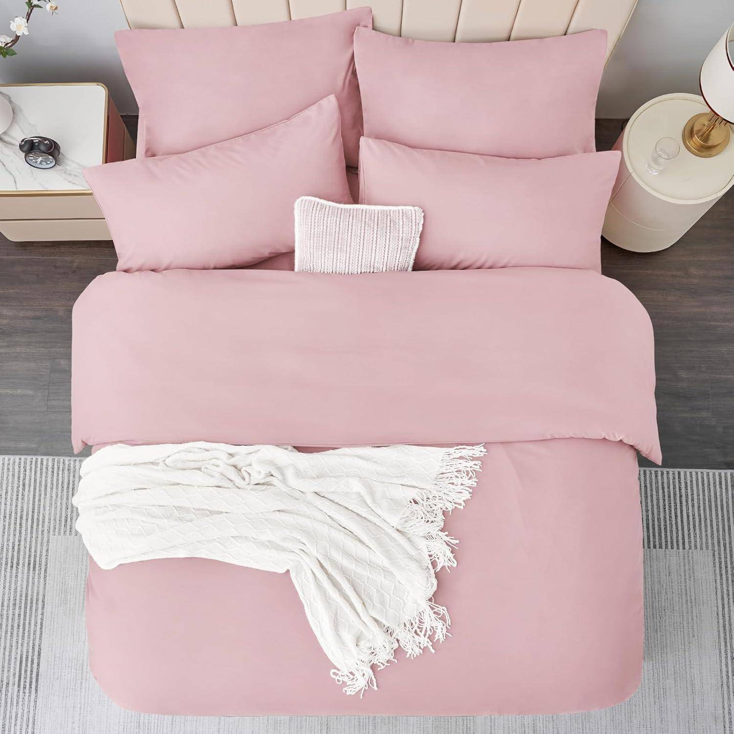 Ein ordentlich hergerichtetes Bett mit rosa Kissen und Bettwäsche, abgerundet durch eine cremefarbene Decke. Ein Nachttisch trägt eine Lampe und Dekoration.