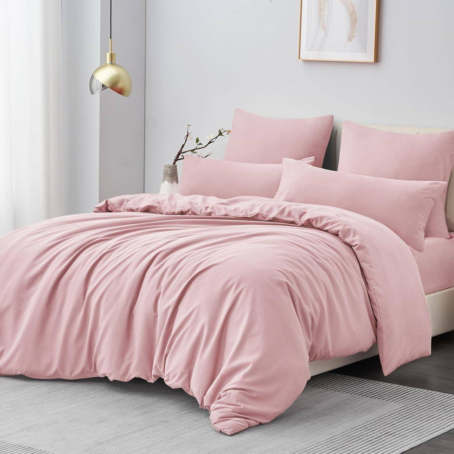 Ein ordentlich gemachtes Bett mit hellrosa Bettwäsche, einschließlich Bettdecke und Kissen, vor einem weißen Kopfteil und einer weißen Wand mit einer goldenen Lampe und gerahmter Kunst.
