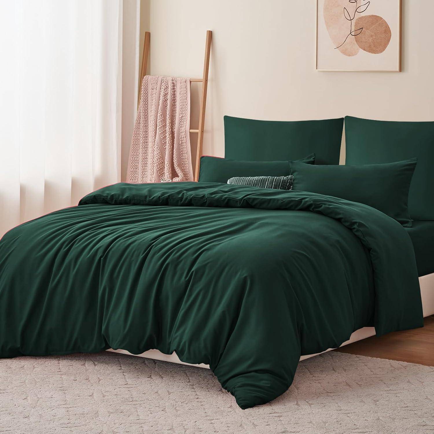 Ein Bett mit dunkelgrüner Bettwäsche und passenden Kissen steht an einer Wand. Eine Leiter mit einer rosa Decke lehnt rechts daneben.