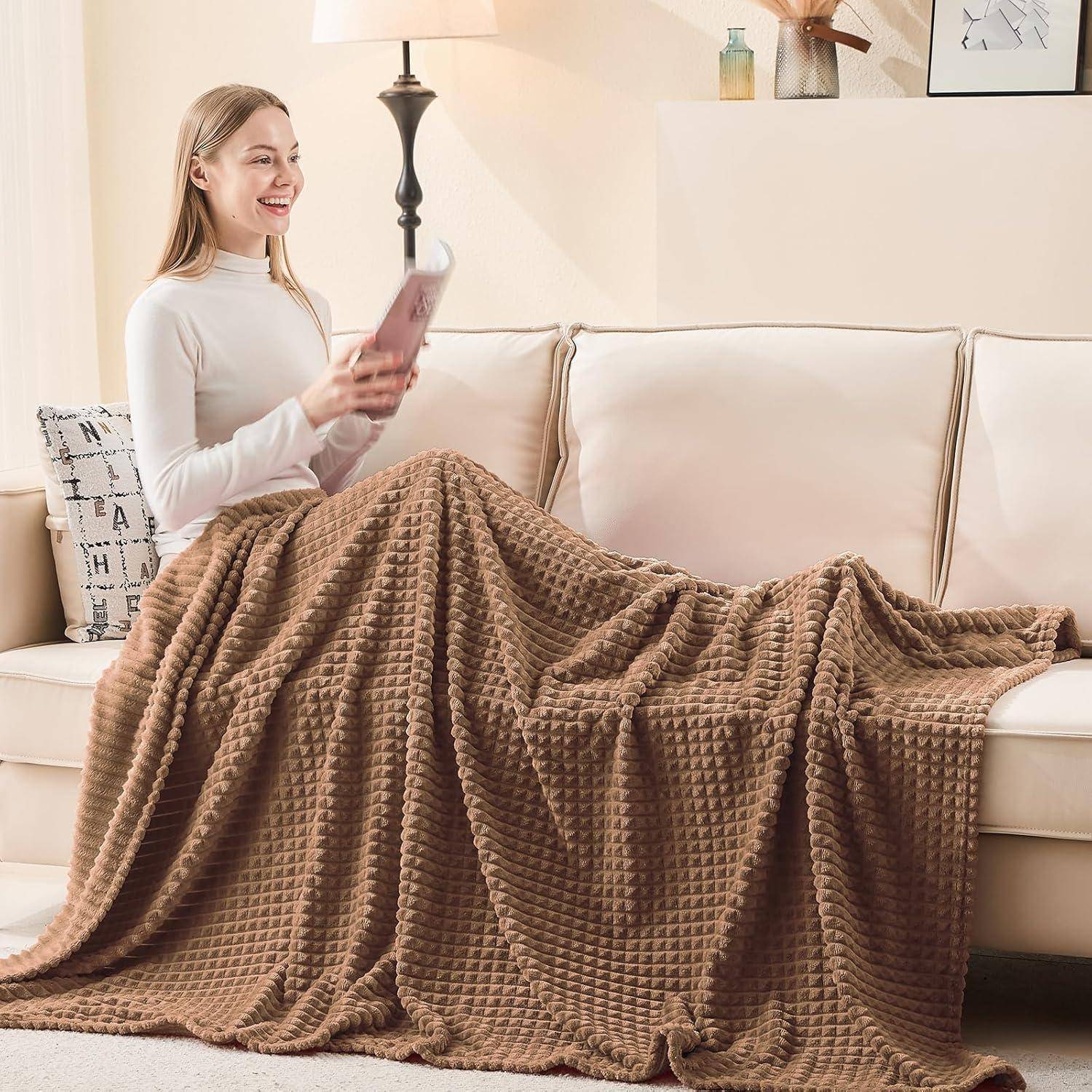 Eine lächelnde Person sitzt auf einem beigen Sofa, das mit einer strukturierten braunen Decke bedeckt ist, hält ein Tablet, neben einer Lampe und einem dekorierten Regal.