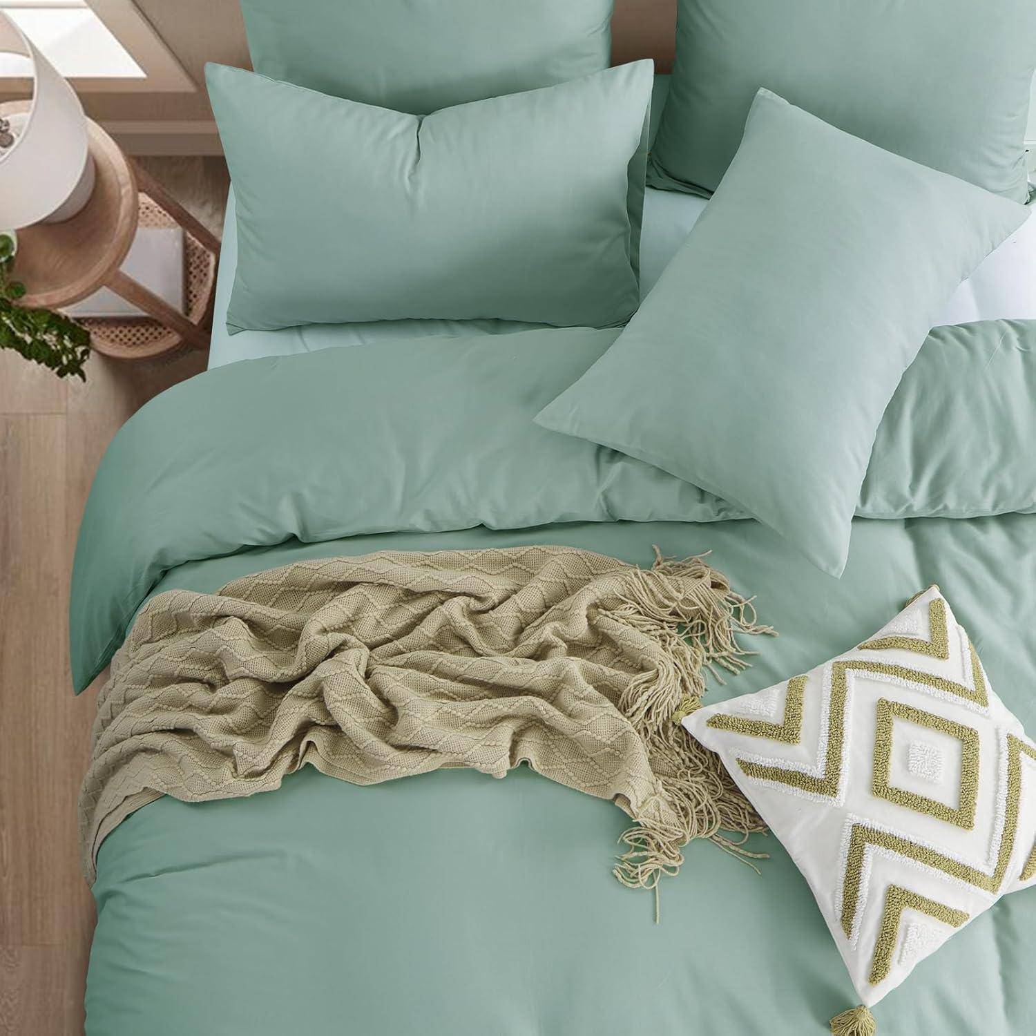Ein ordentlich gemachtes Bett mit hellgrüner Bettwäsche und passenden Kissen, einer beigen gestrickten Decke und einem dekorativen weißen Kissen mit grünen Mustern.