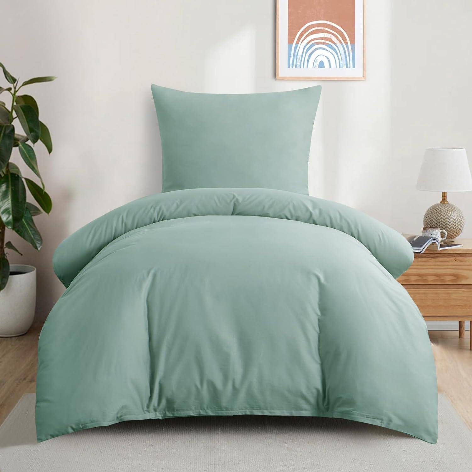 Ein ordentlich gemachtes Bett mit einer hellgrünen Bettdecke und einem Kissen, neben einem Holznachttisch mit einer Lampe. Eine Topfpflanze steht in der Ecke.