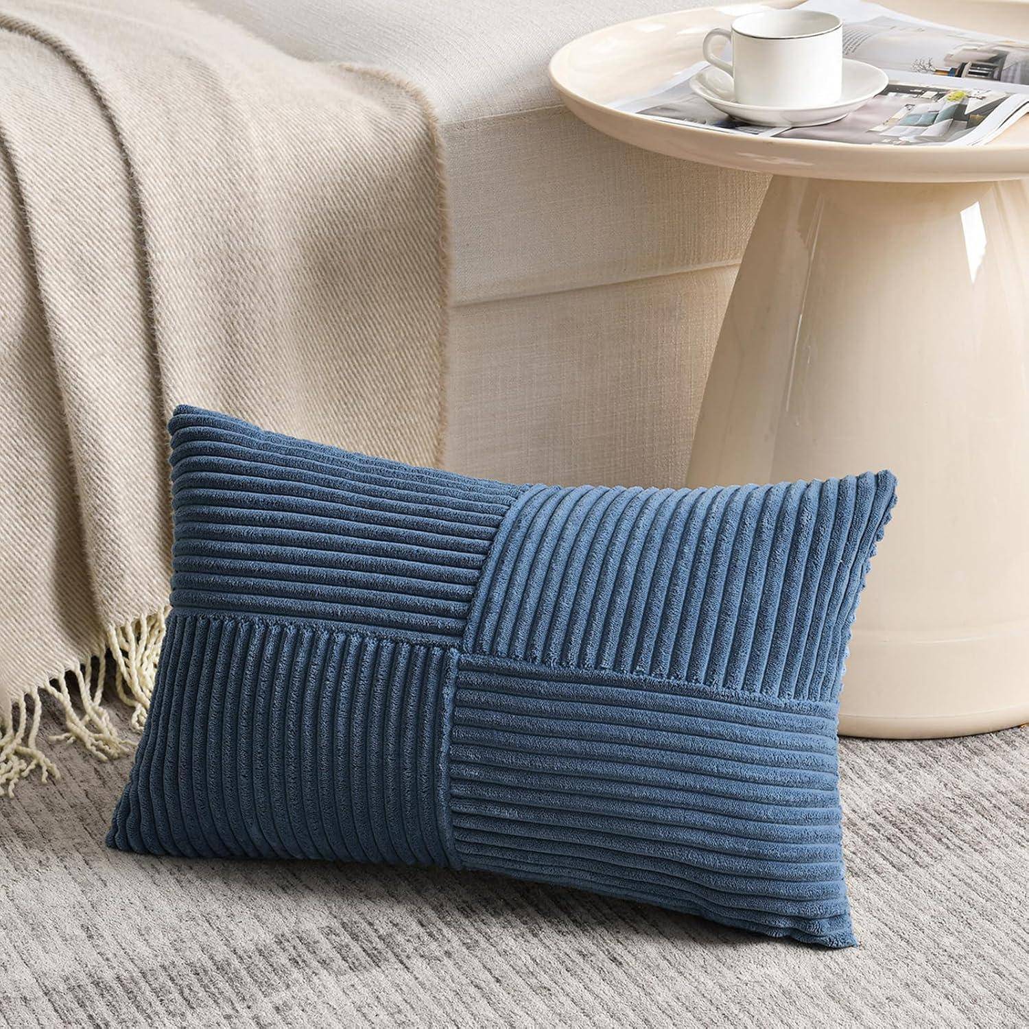 Blauer Cordsamtkissen auf einem beigen Teppich, neben einem weißen Beistelltisch mit einer Tasse und Untertasse, unter einer leichten Decke auf einem Sofa.
