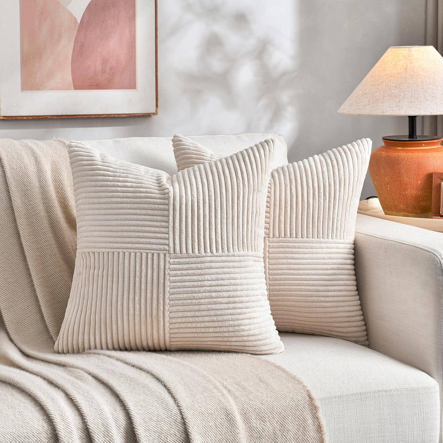 Eine gemütliche Wohnzimmerszene zeigt ein beiges Sofa mit zwei gestreiften Kissen und einer weichen Decke. Eine Lampe und abstrakte Kunstwerke vervollständigen die Einrichtung.