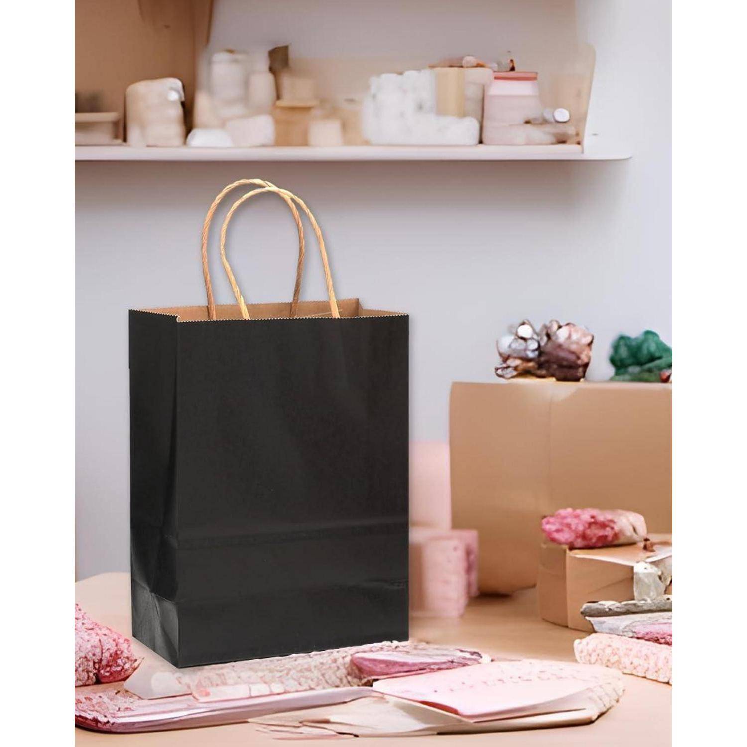 Eine schwarze Geschenktüte aus Papier mit Kordelhenkeln steht auf einem unordentlichen Schreibtisch mit Briefpapier, rosa Seidenpapier und Schachteln in einem Raum mit Regalen.