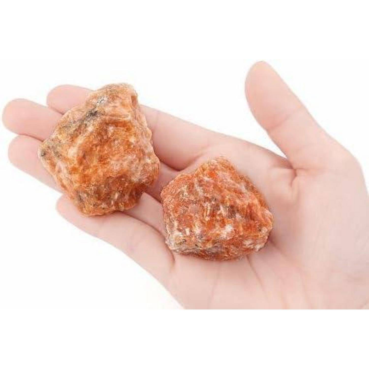 Eine Hand, die zwei raue orangefarbene Kristalle, möglicherweise Mineralien oder Steine, vor einem einfarbigen weißen Hintergrund hält.