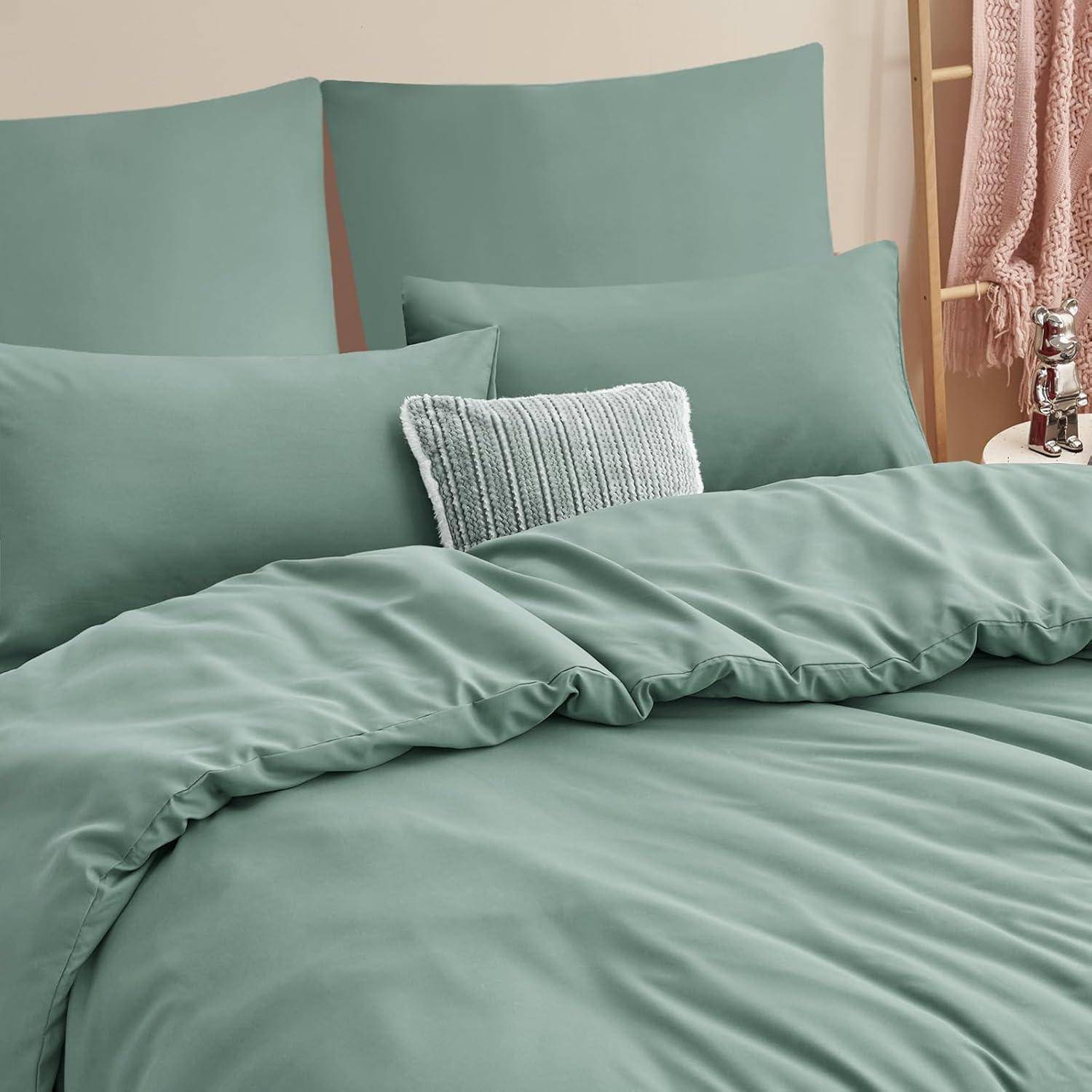 Ein ordentlich gemachtes Bett mit petrolblauer Bettwäsche und zwei Kissen, abgerundet durch ein dekoratives gestricktes Zierkissen. Das Bett steht an einer hellgefärbten Wand.