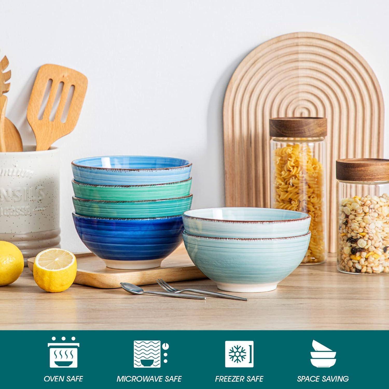 Bunte Keramikschalen auf einer Küchenarbeitsplatte neben Küchenutensilien, Zitronen und Gläsern mit Pasta und Getreide. Text weist darauf hin, dass sie ofenfest, mikrowellengeeignet und gefrierschrankfest sind.