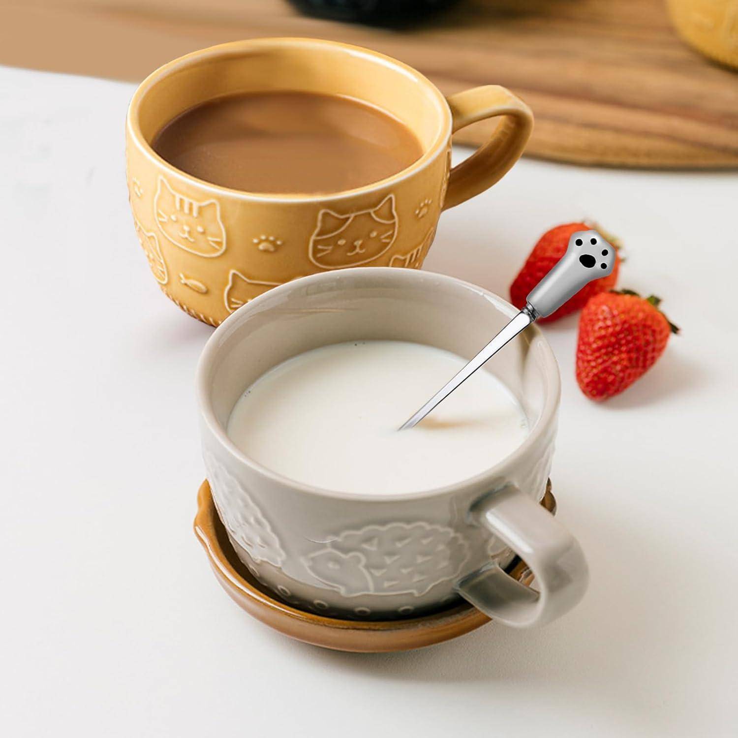 Zwei Keramiktassen auf einem Tisch, eine mit Kaffee, die andere mit Milch. Ein Löffel mit einem Pfotenheft liegt in der Milch. Zwei Erdbeeren daneben.