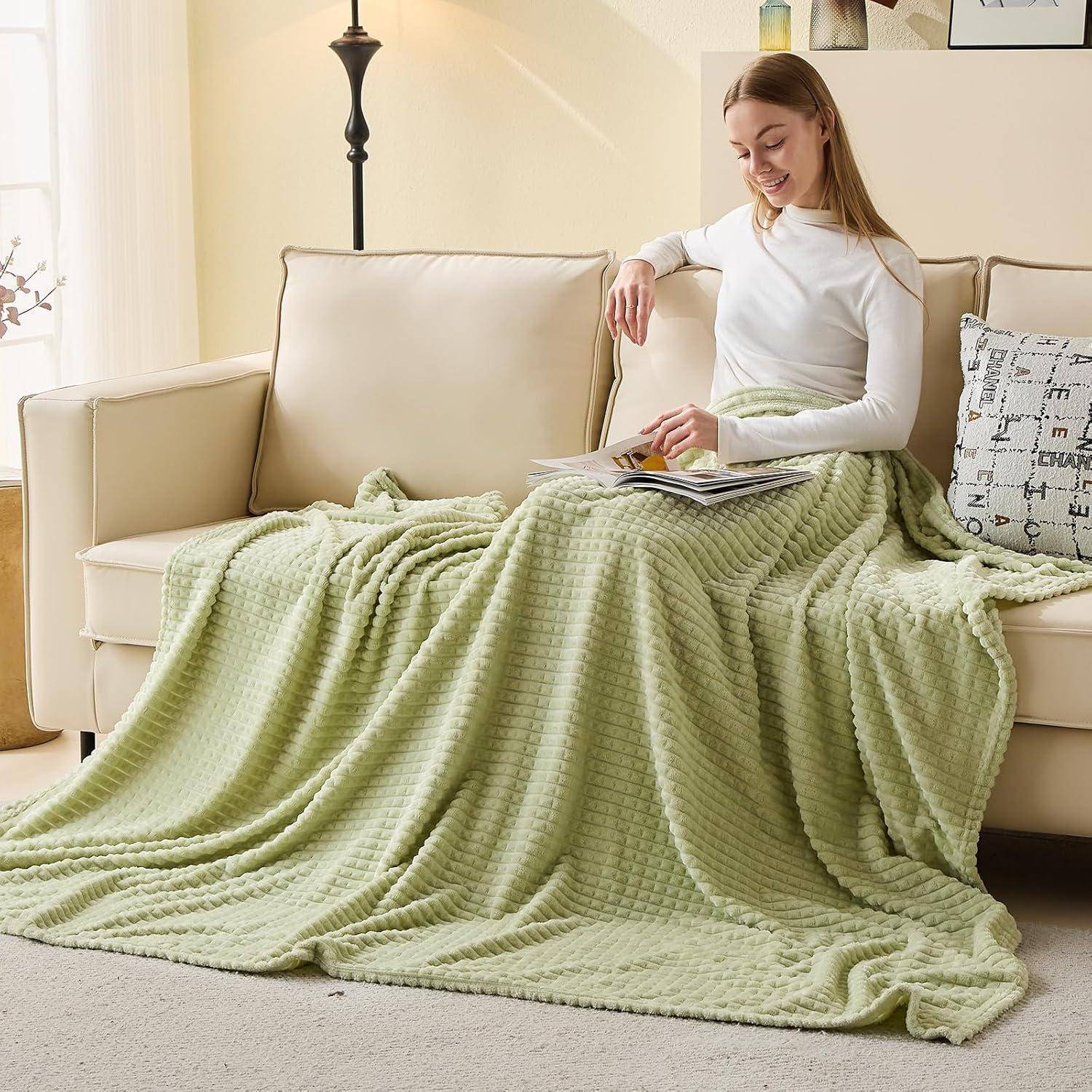 Eine Person, die auf einem beigen Sofa sitzt, mit einer grün gemusterten Decke bedeckt, und ein Buch liest. Warme und gemütliche Wohnzimmeratmosphäre.