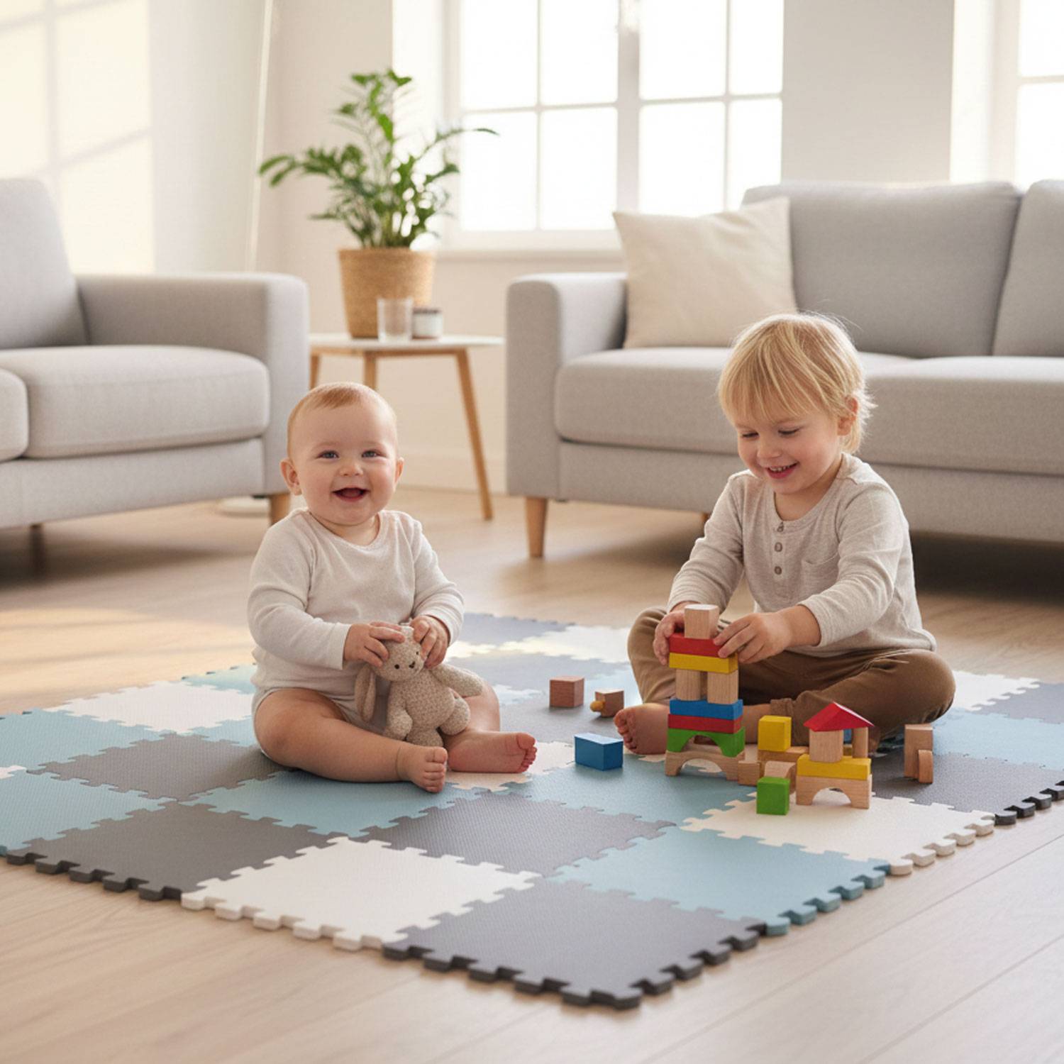 Zwei Kleinkinder spielen auf einer Schaumstoffmatte mit Holzklötzen. Eines baut einen Turm, während das andere lächelt und einen Stofftieridra hält. Gemütliche Zimmeratmosphäre.
