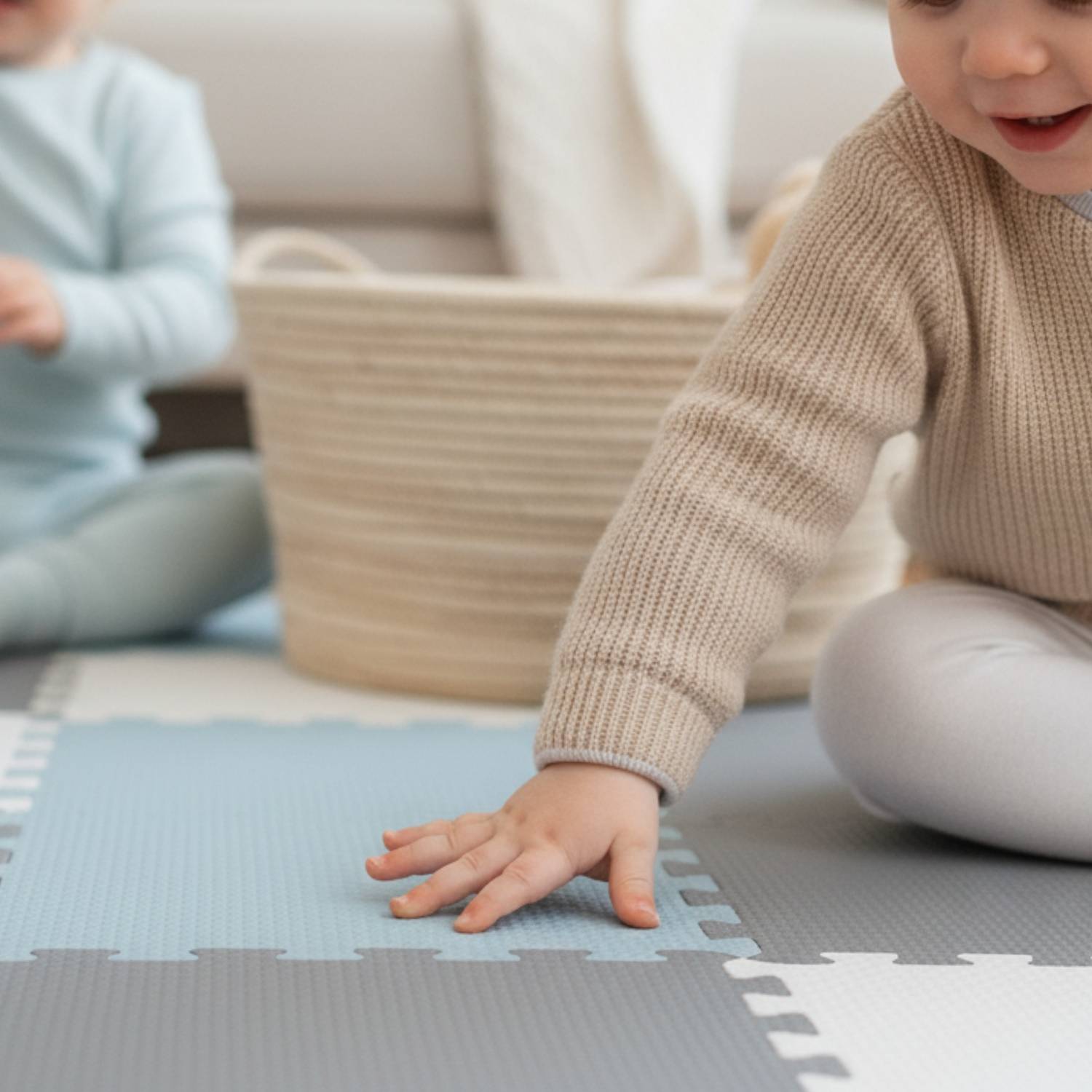 Ein Baby auf einer Schaumstoff-Spielmatte, das einen beigen Pullover trägt, greift nach einem Spielzeug. Ein Weidenkorb und ein weiteres Kind in hellblau sind zu sehen.