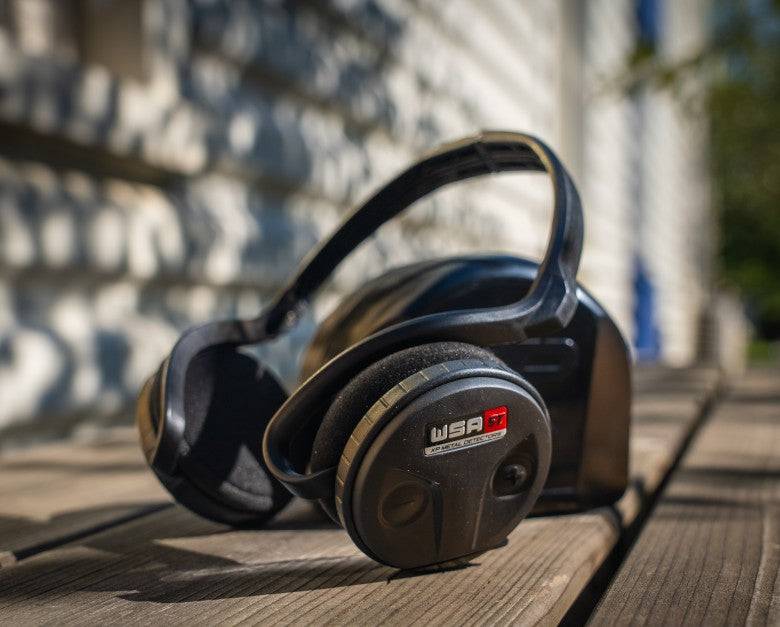 Ein Paar schwarzer Kopfhörer ruht auf einer Holzoberfläche im Freien, mit einem unscharfen Hintergrund aus einer Wand und einigen Blättern.