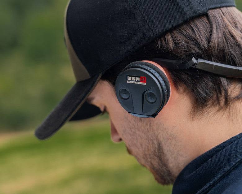 Eine Person mit schwarzen Kopfhörern und einer schwarzen Mütze steht im Freien mit einem unscharfen grünen Hintergrund.