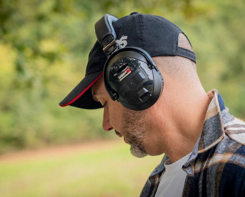 Nahaufnahme eines Mannes, der draußen Gehörschutz-Ohrenschützer und eine Kappe trägt, was auf Lärmschutz oder Schießstand-Nutzung hindeutet.